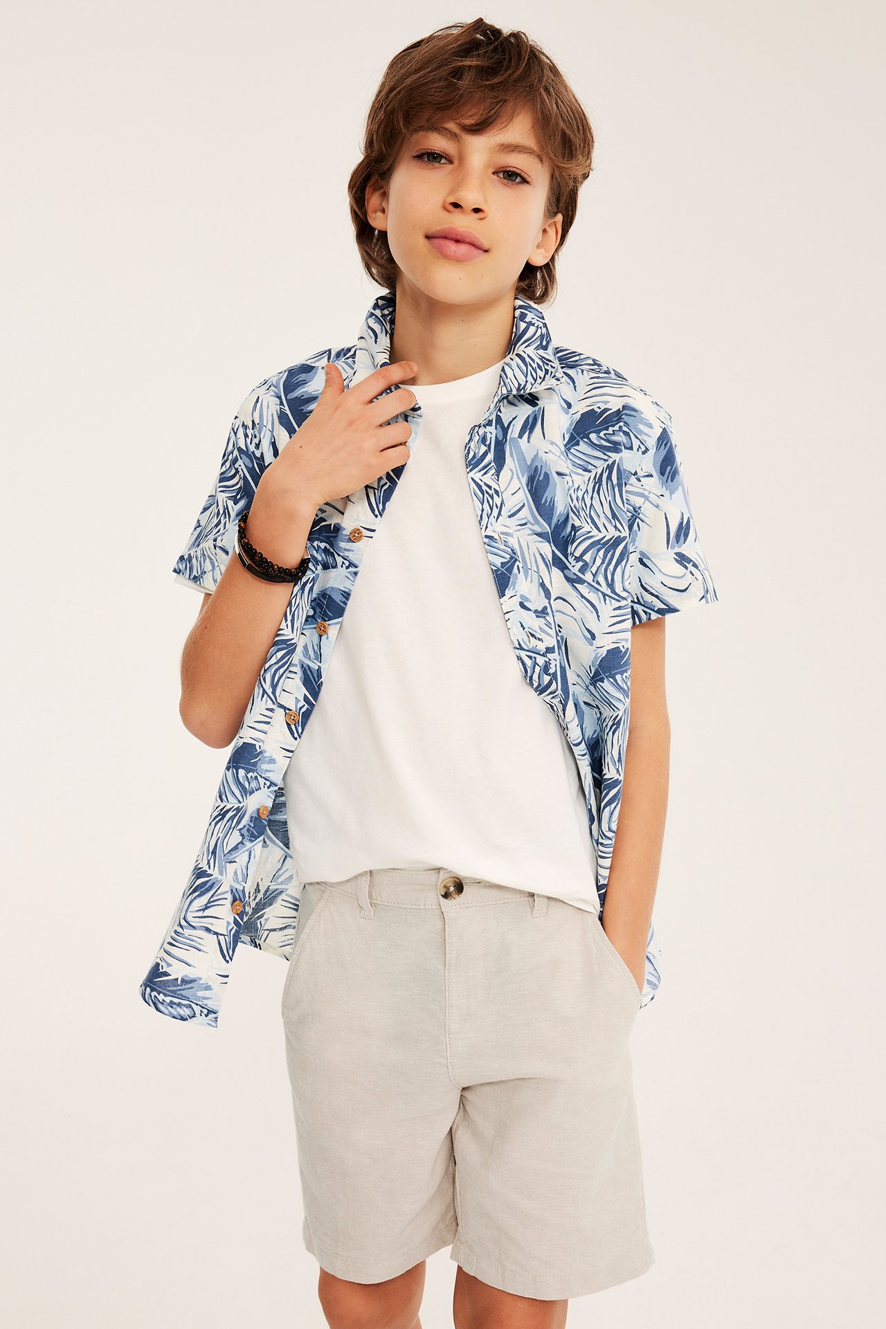 Edestäpäin kuvattu lapsi vaaleabeigeissä pellavasekoiteshortseissa, valkoisessa t-paidassa ja avoimessa sinikuvioisessa paidassa.