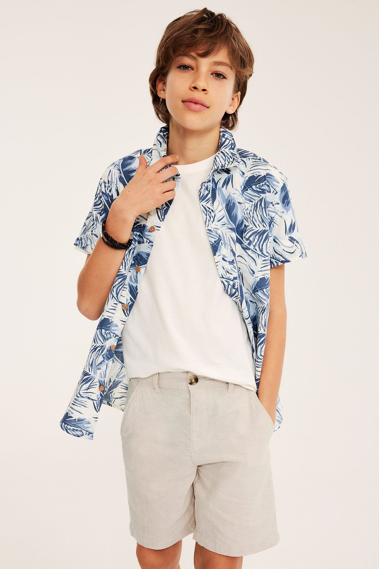 Edestäpäin kuvattu lapsi vaaleabeigeissä pellavasekoiteshortseissa, valkoisessa t-paidassa ja avoimessa sinikuvioisessa paidassa.