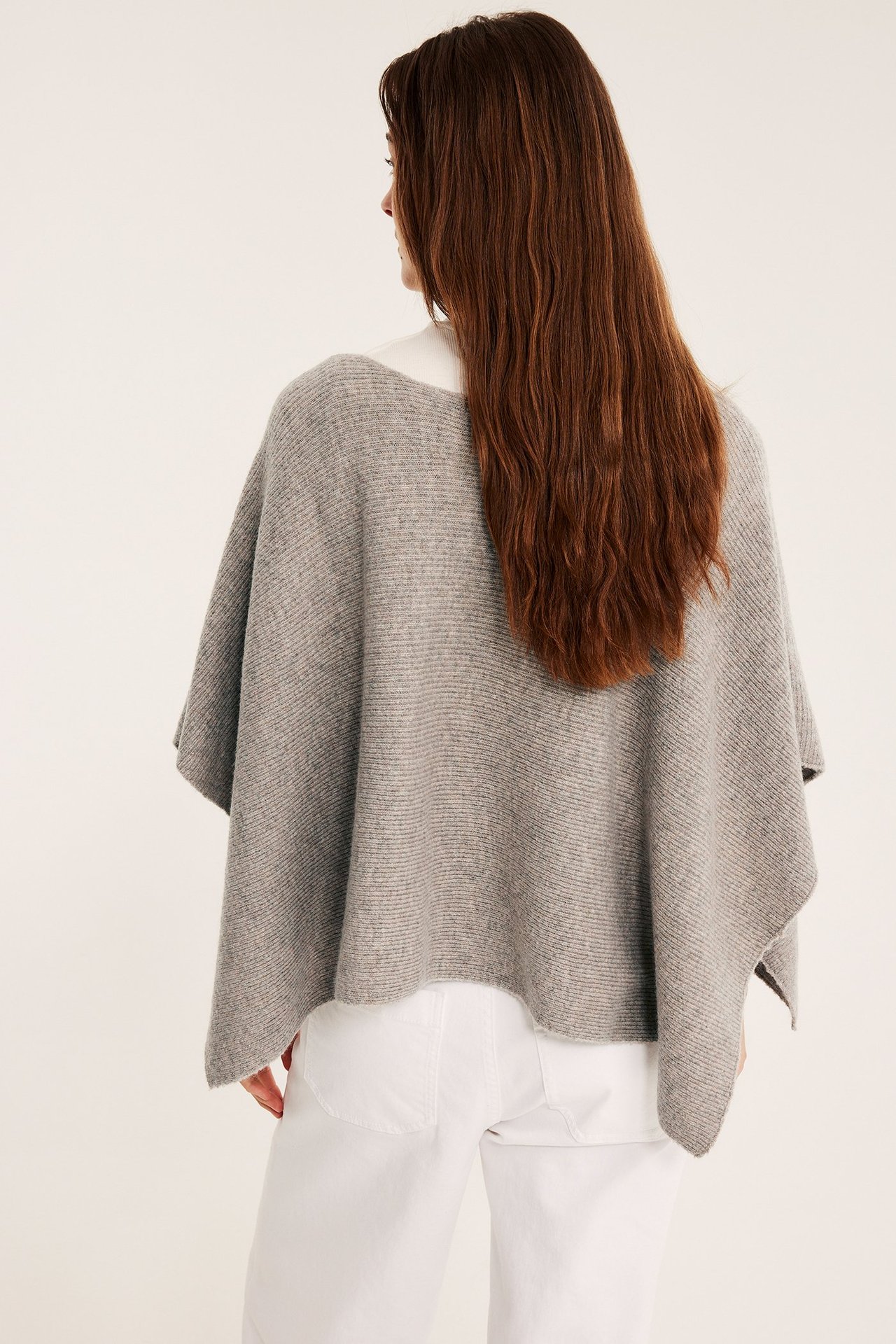 Bakifrån syns en kvinna i en beigemelange stickad poncho över en vit topp och vita byxor.