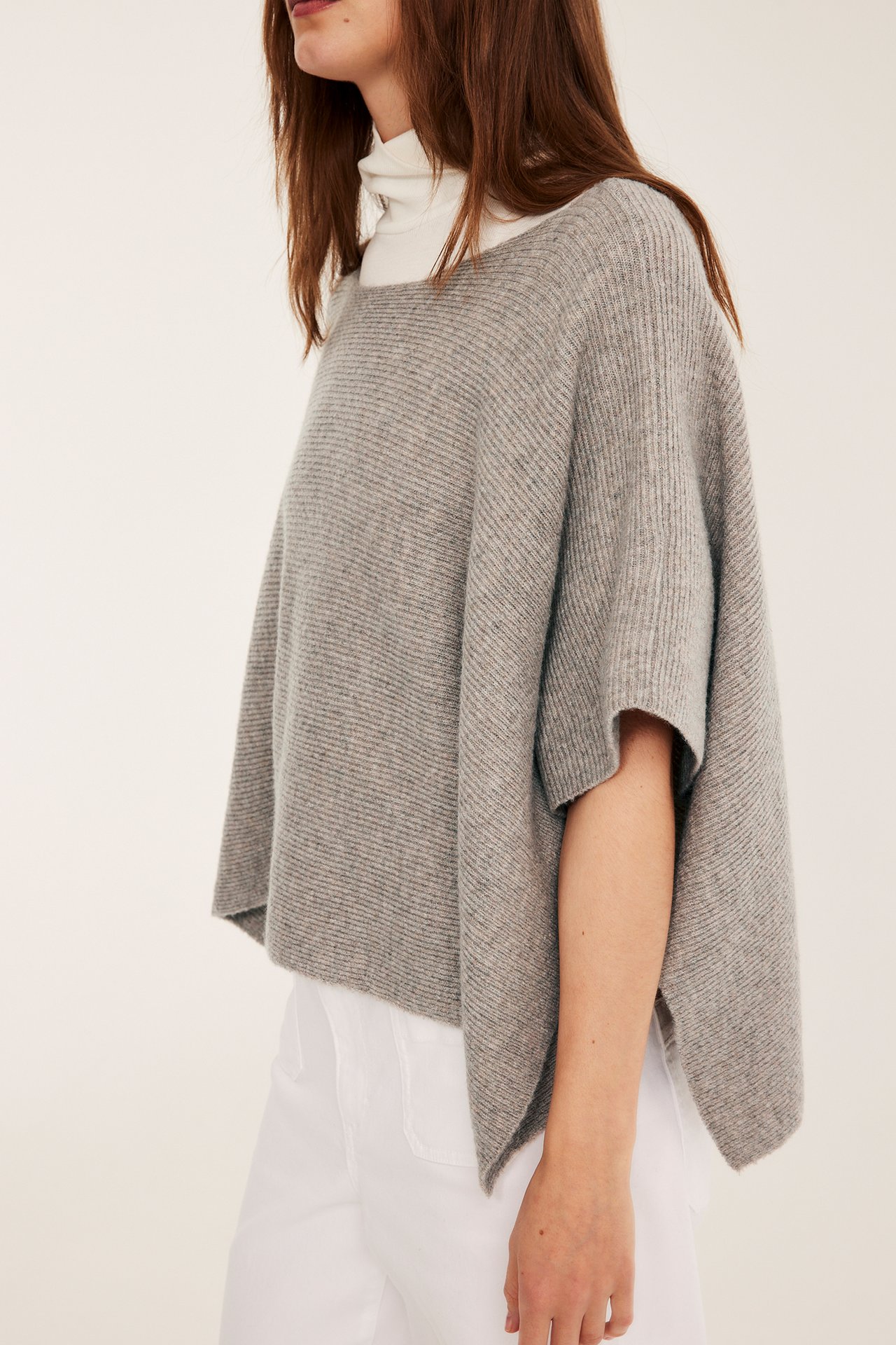 Etunäkymä naisesta, yllään väljä beige melange neulottu venepääntieponcho valkoisen poolopaidan päällä.