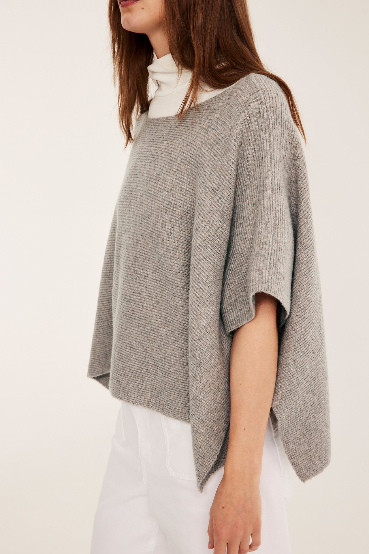 Frontbilde av en kvinne i en løs strikket båthalsponcho i beige melange over en hvit pologenser.