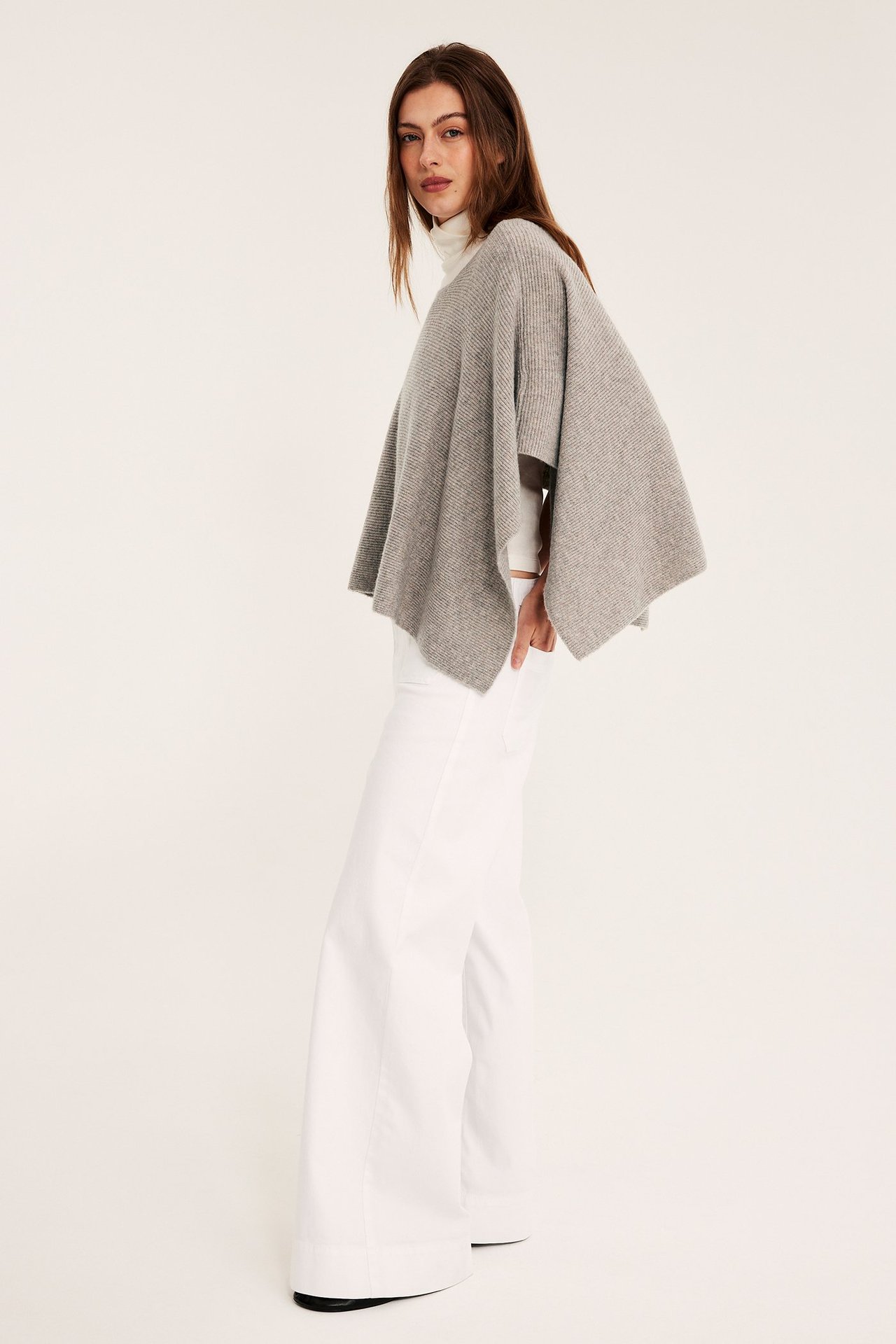 Sideutsikt: kvinne i en melert beige strikket poncho, hvit høyhalset topp og hvite vide bukser.