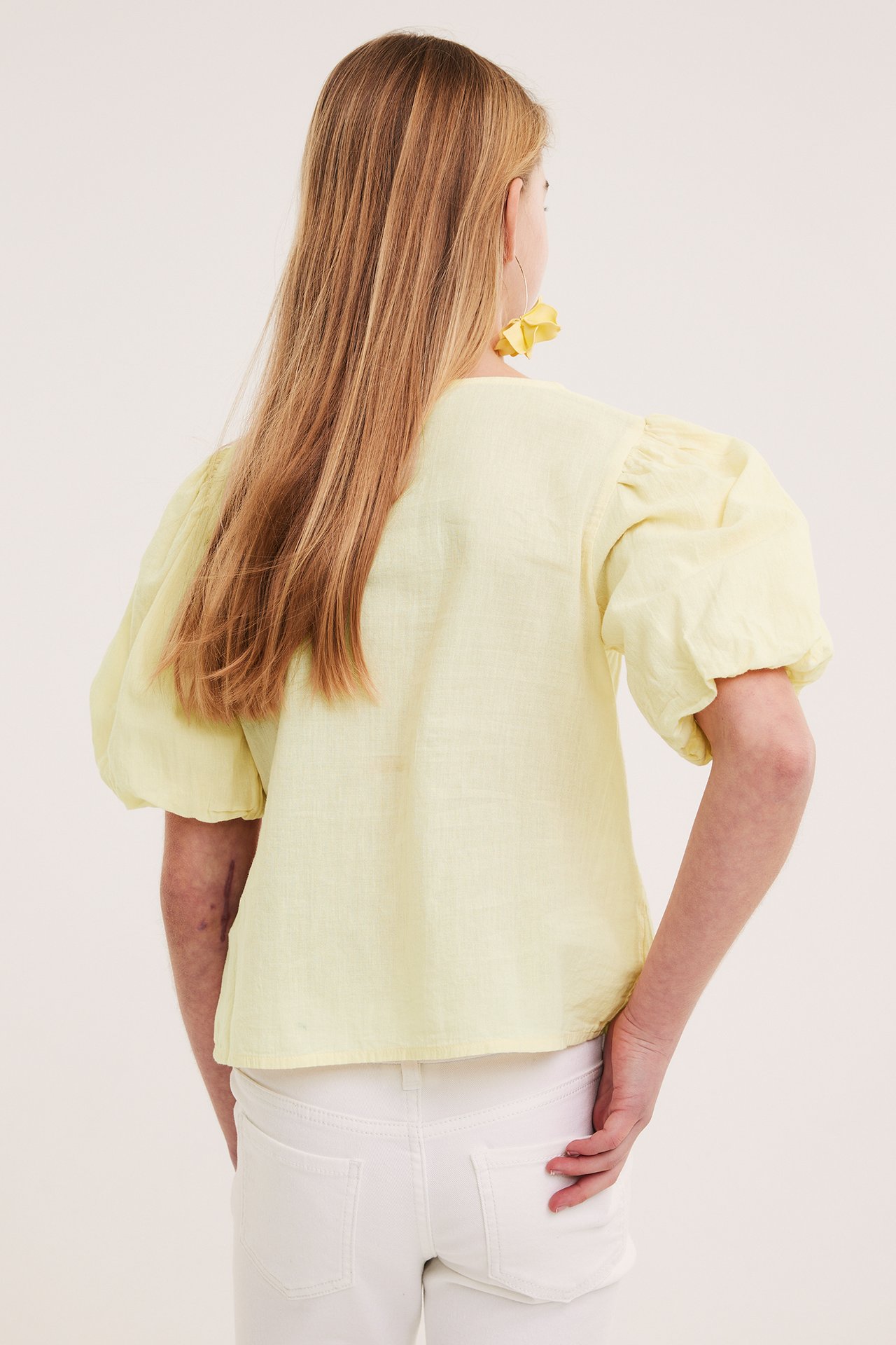 Ryggvy av ett barn i ljusgul puffärmsblus, vita jeans och gula blommiga örhängen.