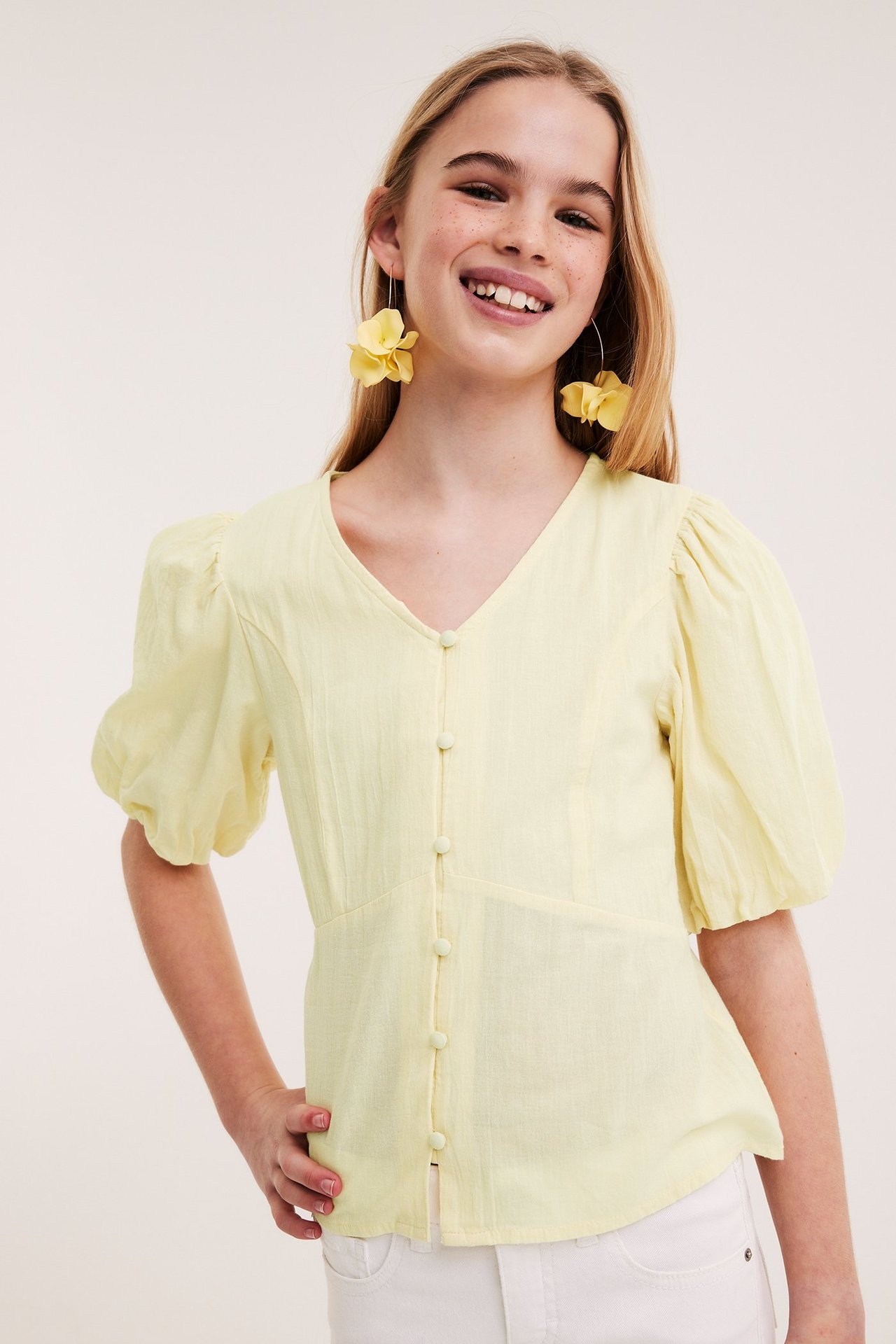 Framsida: Tjej i ljusgul blus med puffärm och knappar, vita byxor och gula blomörhängen.