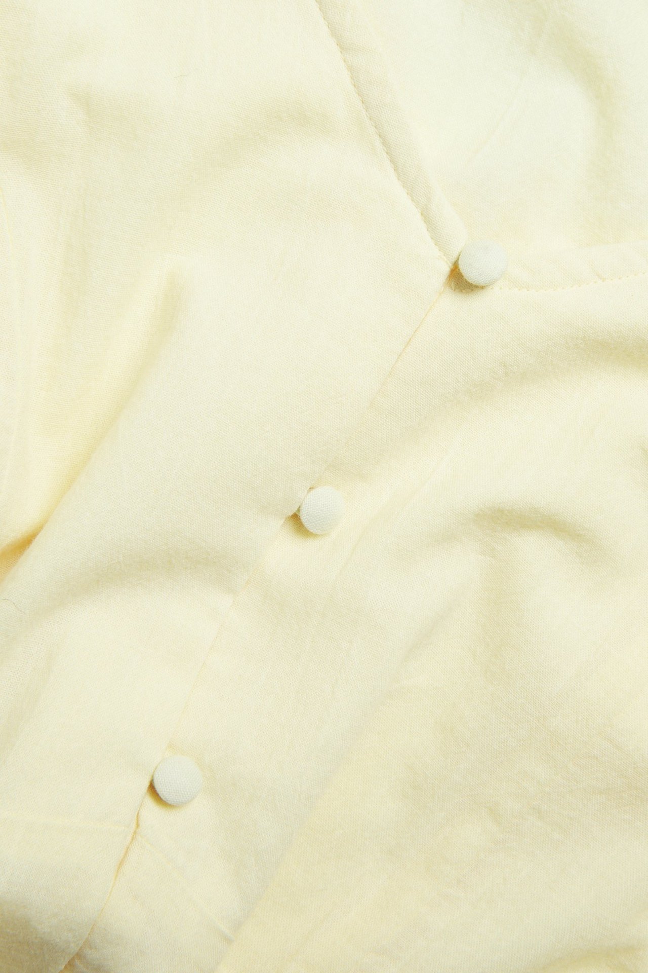 Zbliżenie przodu jasnej, żółtej, jednokolorowej bluzki z trzema obciąganymi guzikami.