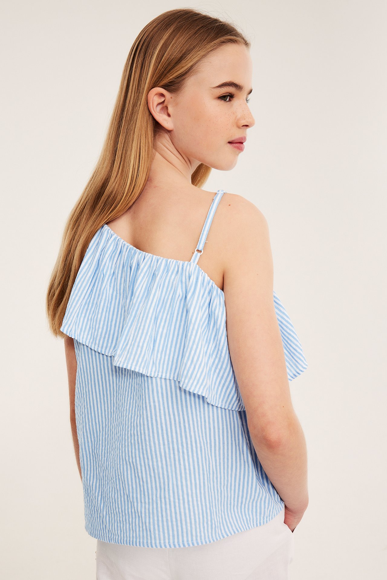 Widok z boku dziecka w jasnoniebieskim topie na jedno ramię z falbanką i białych spodniach.