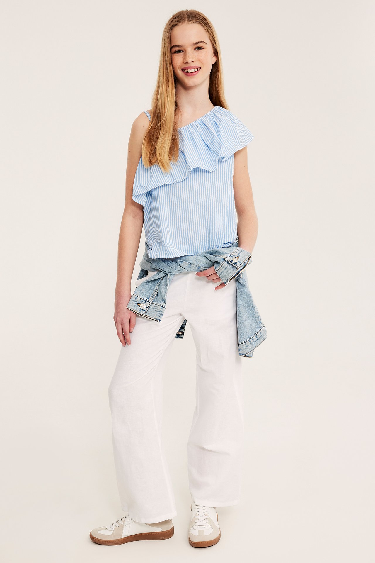 Widok z przodu, dziecko w jasnoniebieskim, pasiastym topie one-shoulder, białych spodniach, kurtce jeansowej i trampkach.