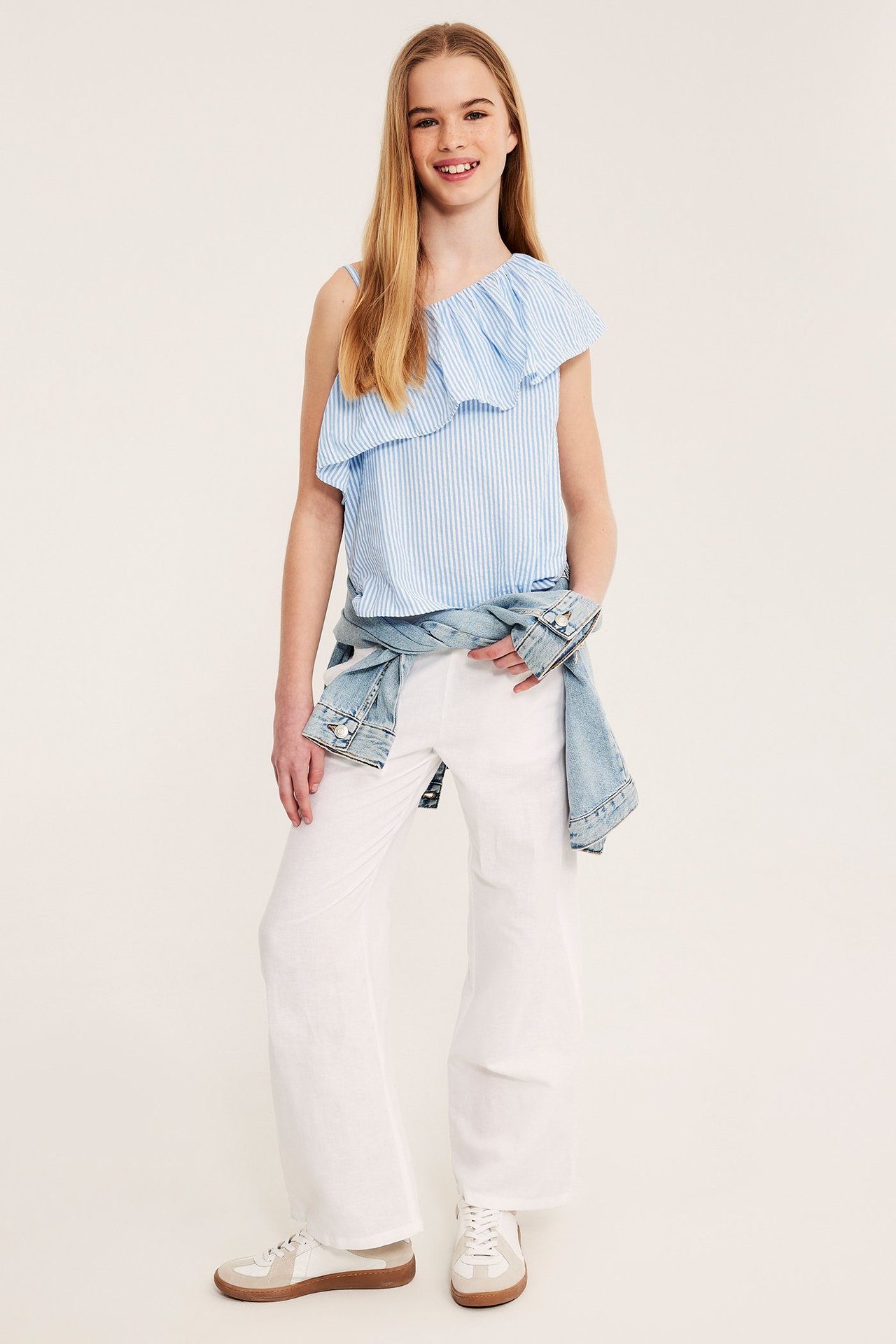 Edestä, lapsi vaaleansinisessä raidallisessa yksiolkaintoppi röyhelöllä, valkoisissa housuissa, farkkutakissa ja lenkkareissa.