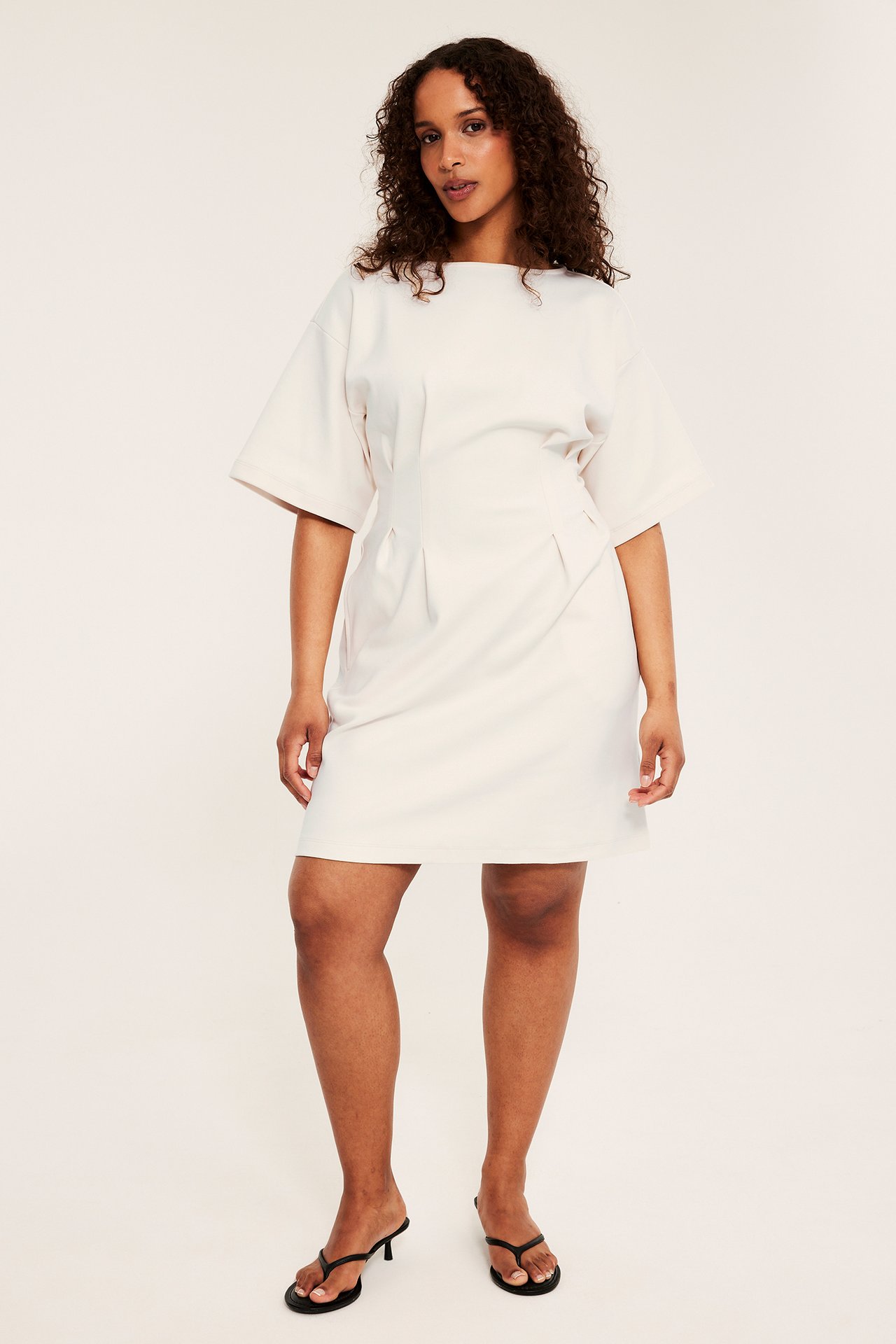 Framifrån, kvinna i offwhite kort jerseyklänning med veck i midjan och svarta högklackade flip-flops.