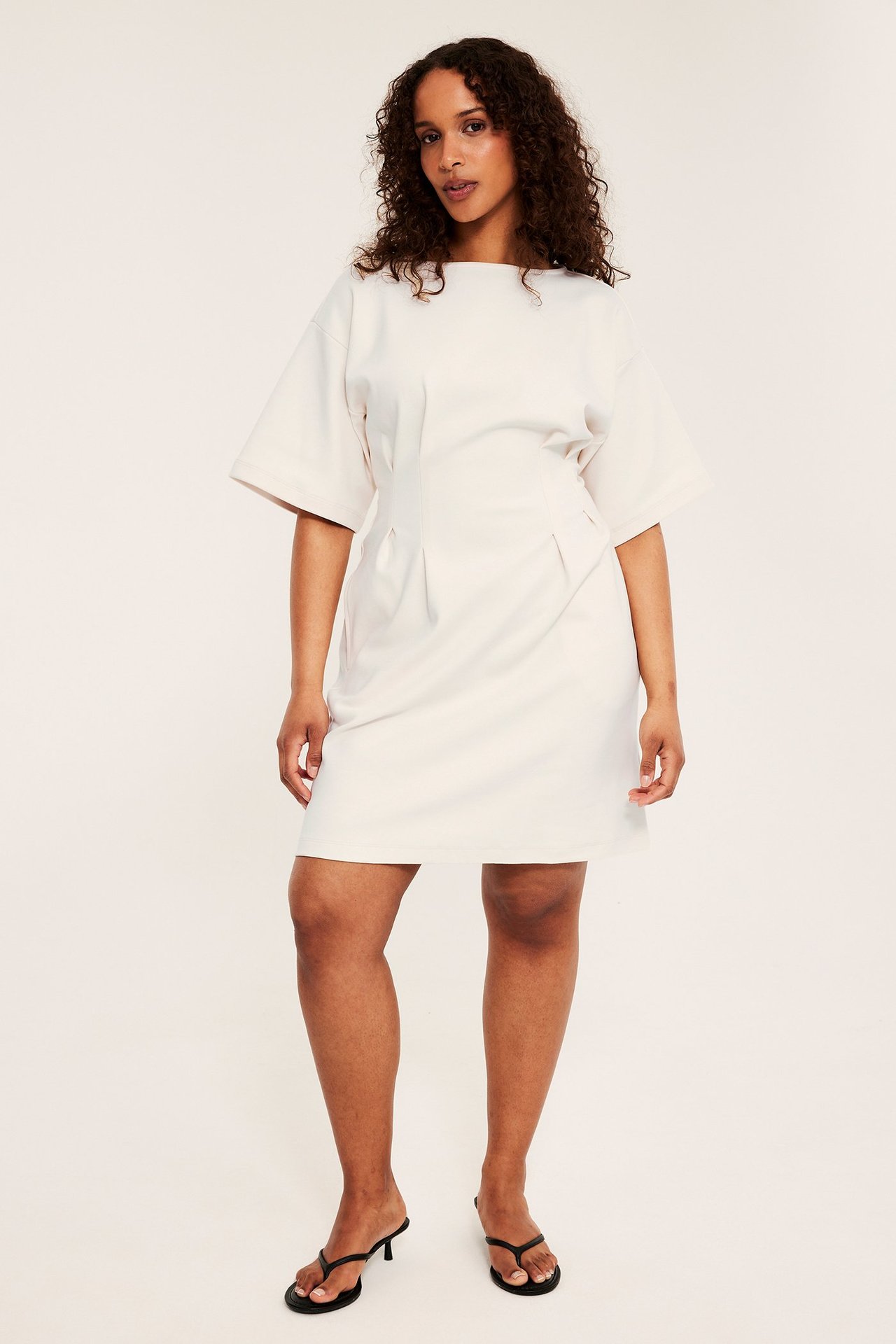 Frontbilde av kvinne i offwhite kort jerseykjole med folder og svarte høyhælte sandaler.