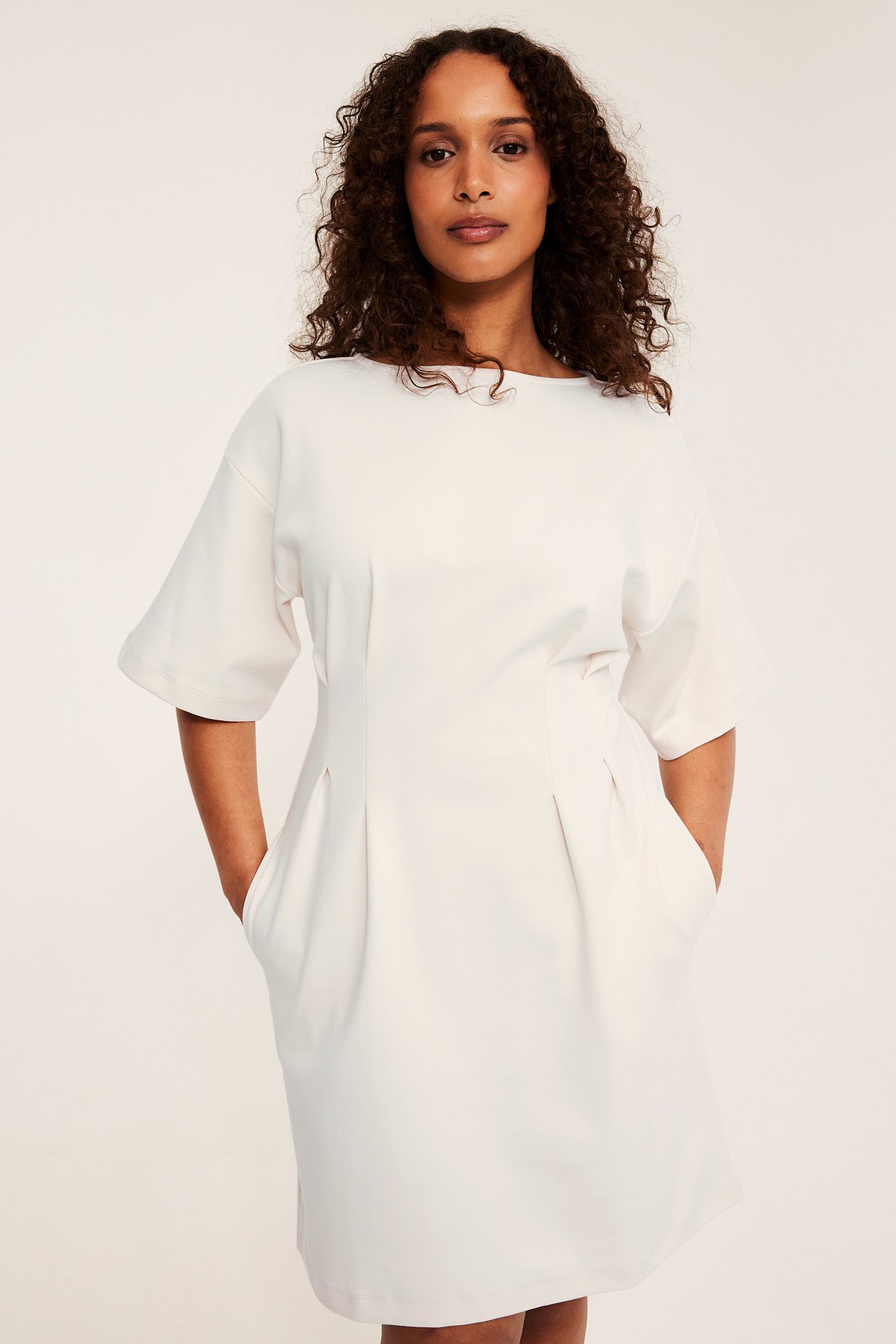 Framifrån, kvinna i offwhite kort jerseyklänning med veck i midjan och fickor.