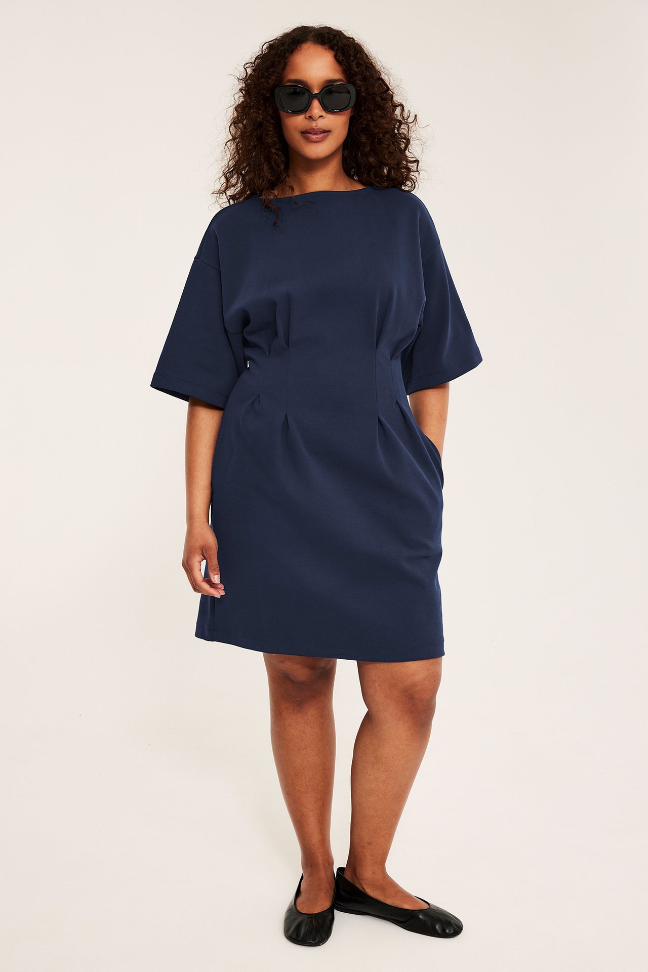 Framifrån en kvinna i mörkblå kort jerseyklänning med veckad midja, svarta ballerinaskor och solglasögon.