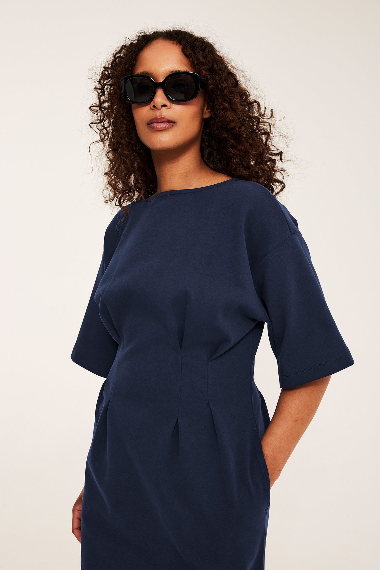Forfra av en kvinne i mørkeblå jerseykjole med folder og svarte rektangulære solbriller.