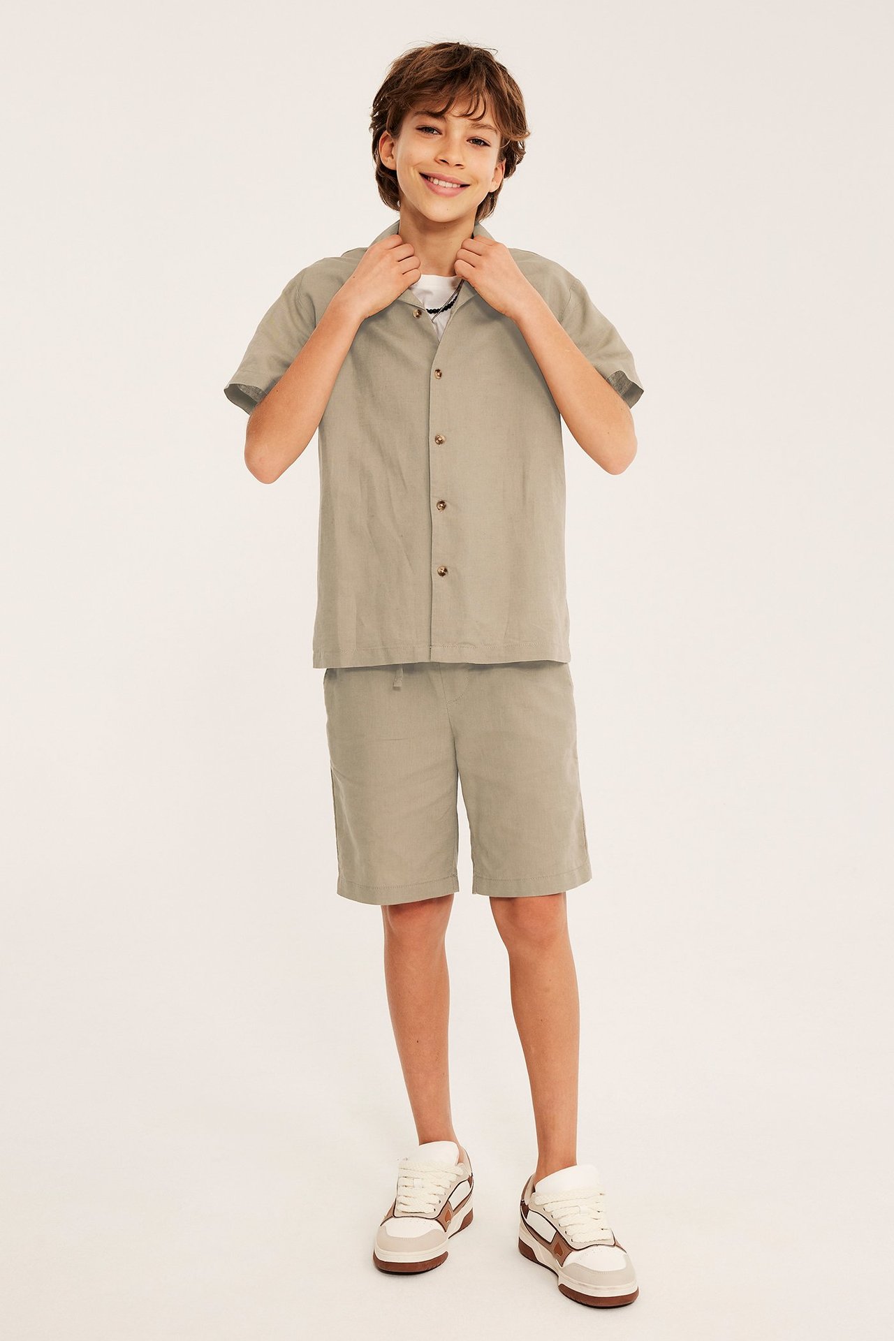 Widok z przodu: Dziecko w beżowych lnianych szortach, pasującej koszuli z krótkim rękawem na białym t-shircie i sneakersach.