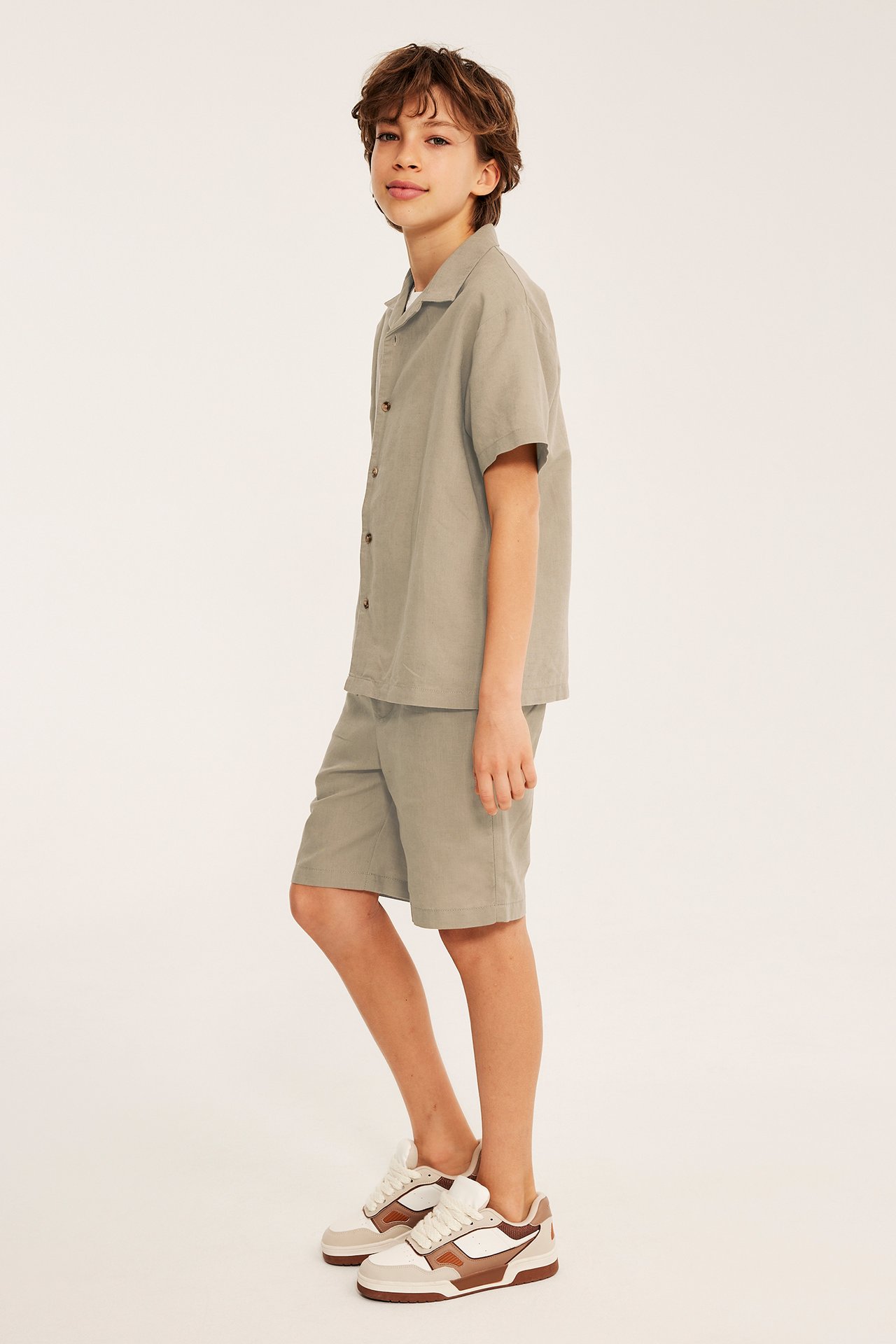 Sidovy av ett barn i beige linne-shorts, matchande kortärmad skjorta och sneakers.