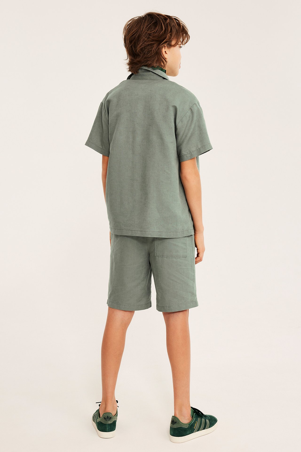 Takakuva: Lapsi vihreissä pellavasekoiteshortseissa, yhteensopivassa lyhythihaisessa paidassa ja vihreissä tennareissa.