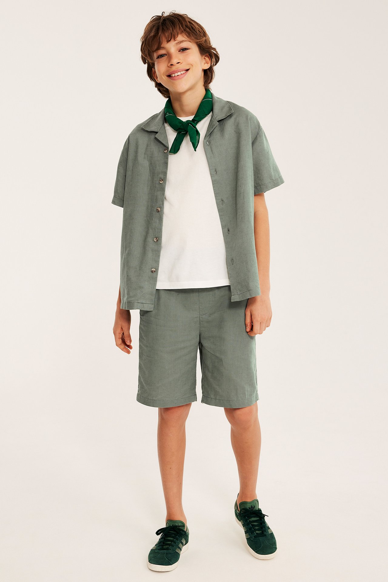 Edestäpäin: Lapsi vihreissä pellavasekoiteshortseissa, yhteensopiva paita valkoisen t-paidan päällä ja vihreät lenkkarit.