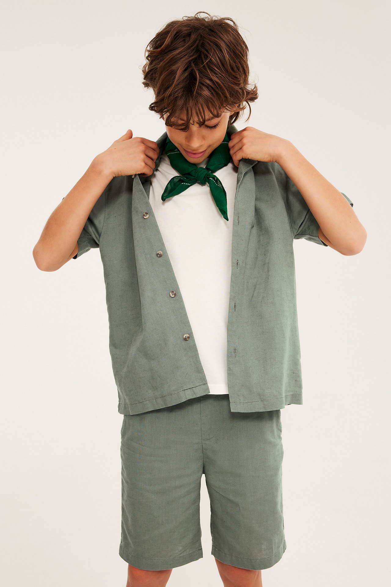 Edestä, lapsi vihreissä pellavasekoiteshortseissa, yhteensopiva avoin paita, valkoinen t-paita ja vihreä huivi.