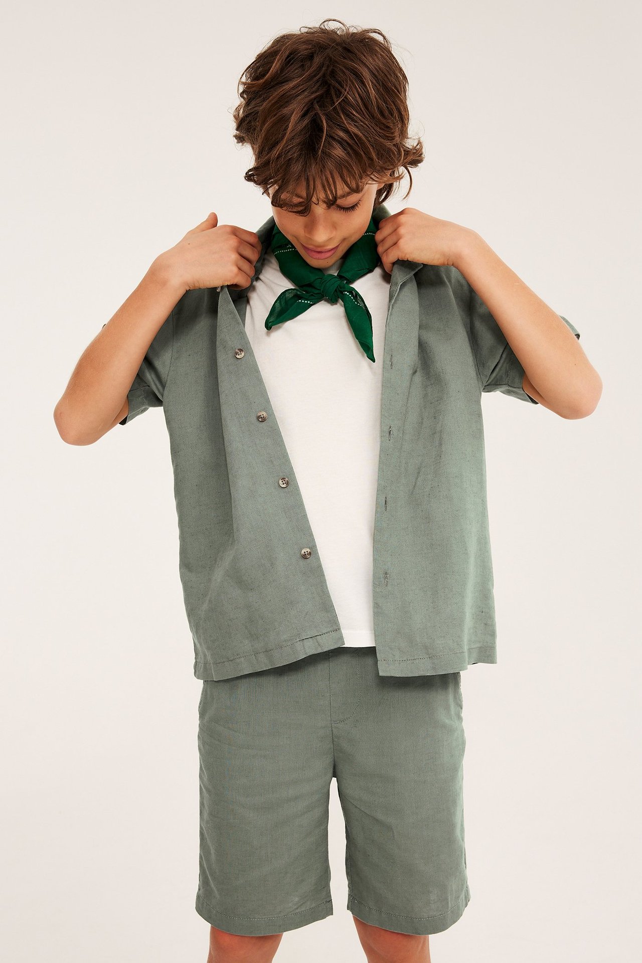 Edestä, lapsi vihreissä pellavasekoiteshortseissa, yhteensopiva avoin paita, valkoinen t-paita ja vihreä huivi.