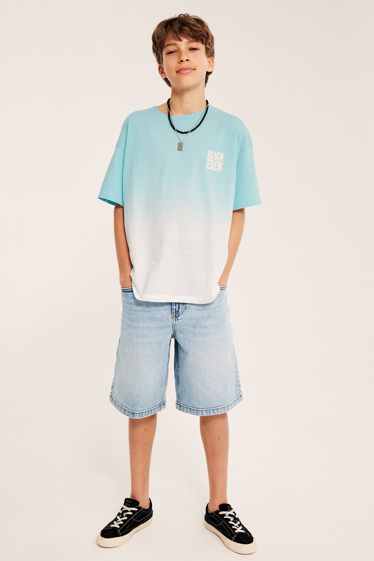 Framifrån: Ett barn i en oversized ljus turkos t-shirt med tryck, ljusa jeansshorts och svarta sneakers.