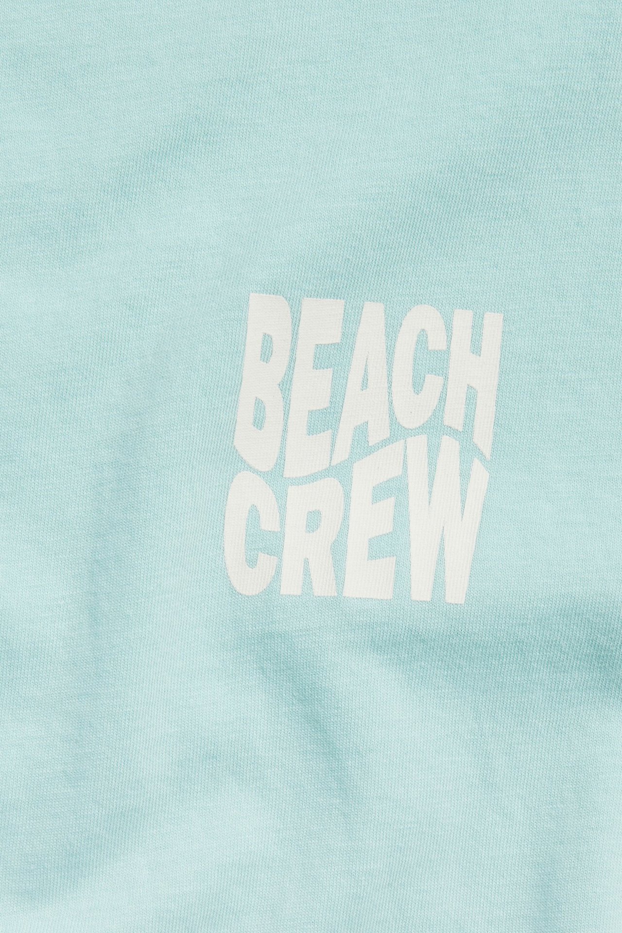 Lähikuva vaalean turkoosista oversize puuvillaisesta t-paidasta valkoisella 'BEACH CREW' etupainatuksella.