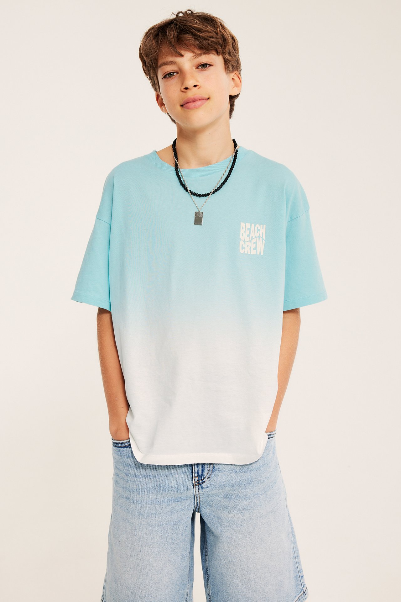 Frontvy: Pojke i oversized ljus turkos-vit ombré T-shirt med tryck, jeansshorts och ett halsband.