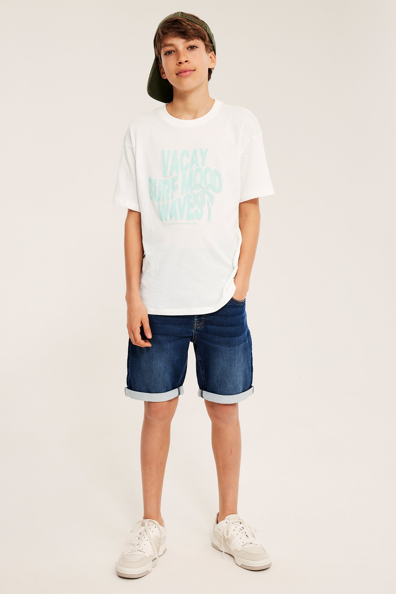 Frontvy: Pojke bär en offwhite oversize t-shirt med tryck, mörkblå shorts, vita sneakers, grön keps.