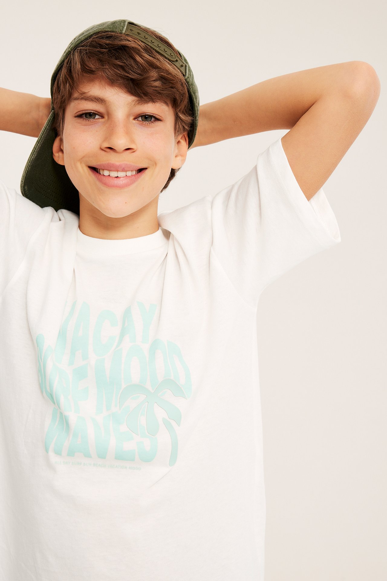 Framifrån: Barn bär offwhite oversize t-shirt med ljusblått tryck och en grön keps.