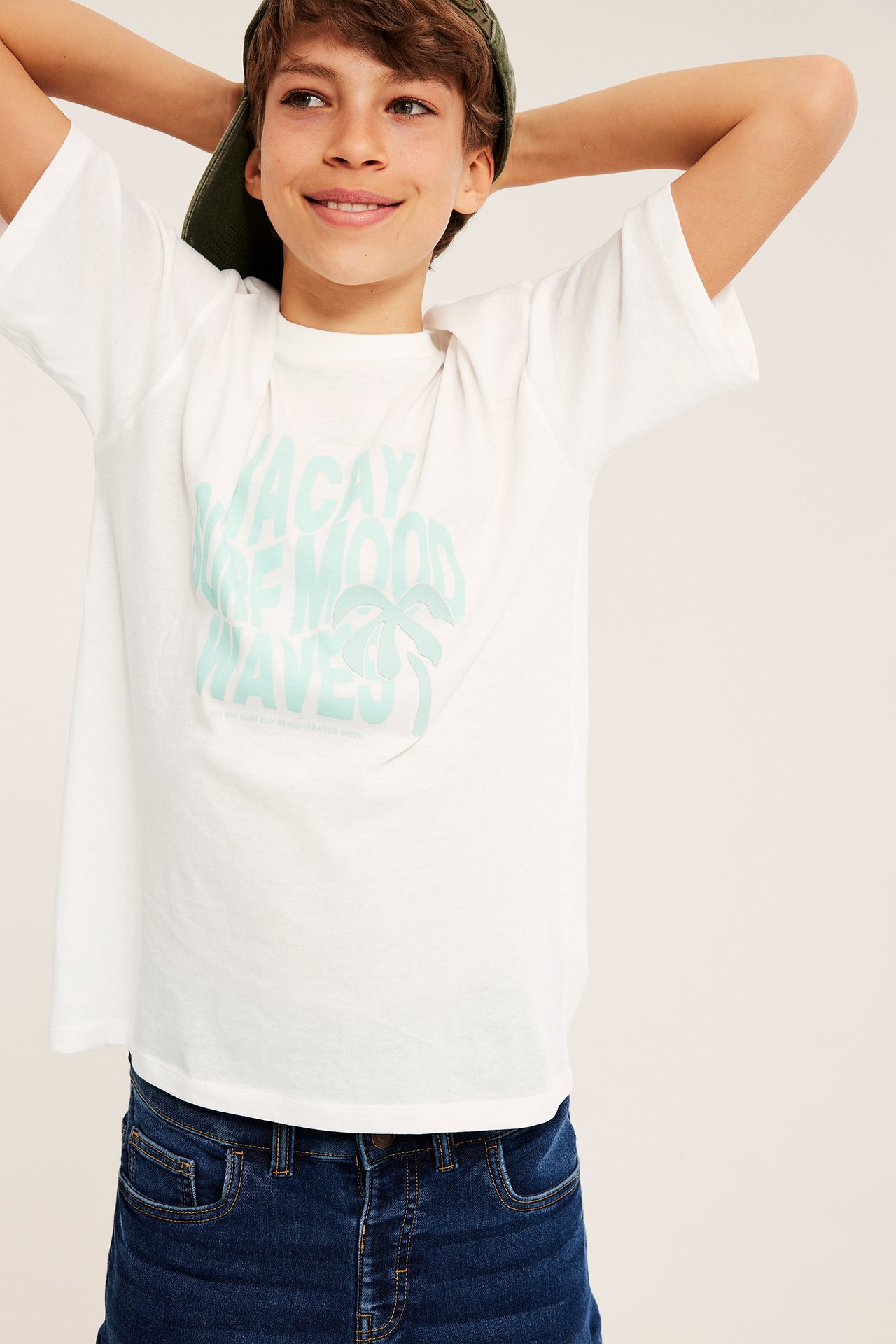 Framifrån: Barn i off-white oversize bomulls-t-shirt med ljusblått tryck, mörkblå jeans, grön keps.