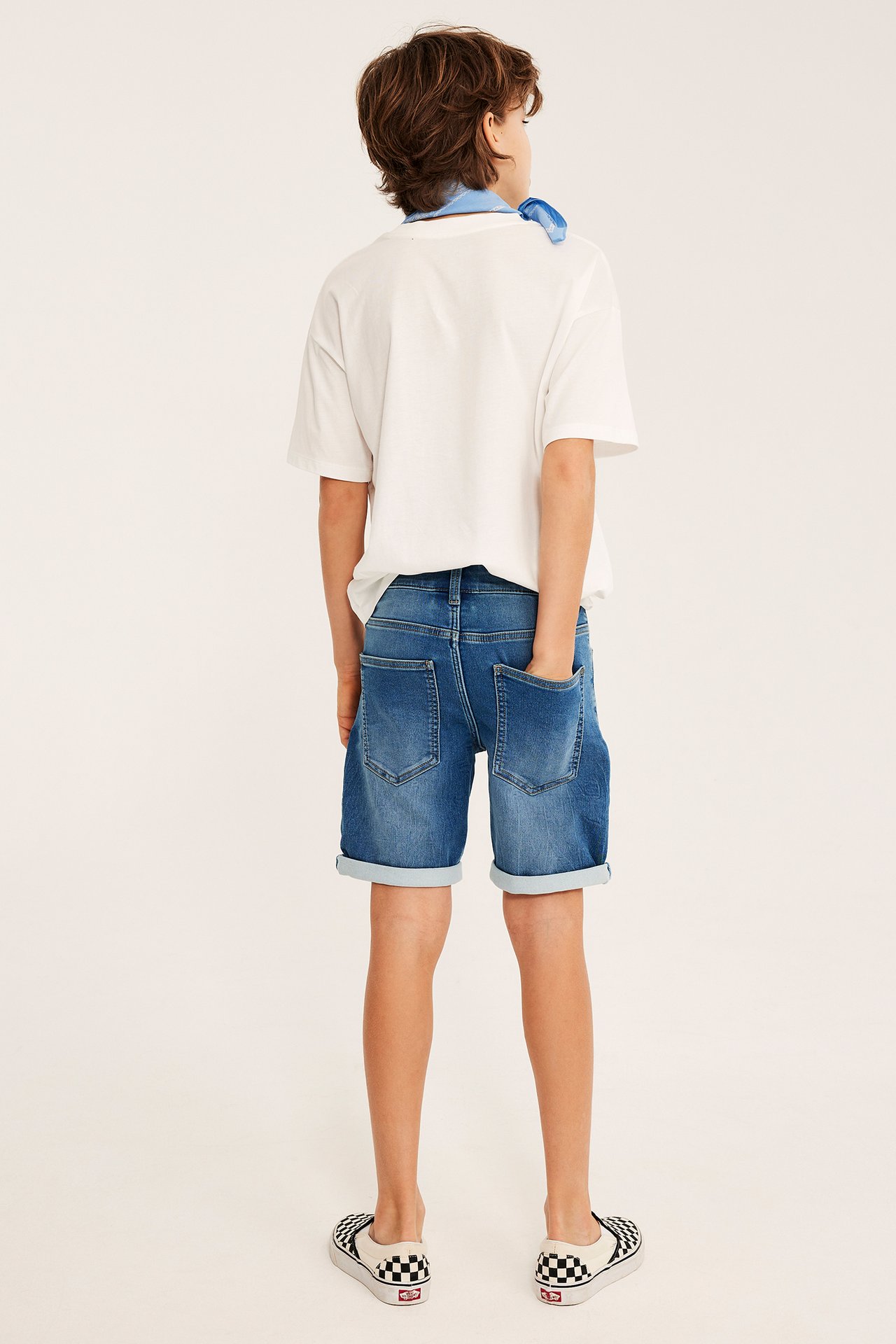 Bakifrån, ett barn i ljusblå joggerjeansshorts, vit t-shirt, blå bandana och rutiga skor.