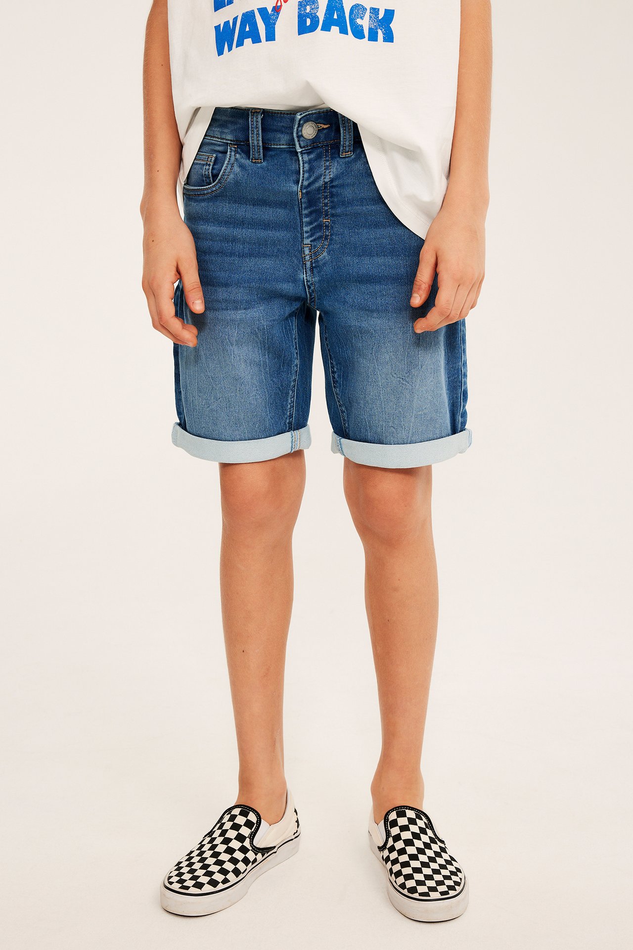 Framifrån, ett barn i ljus denim jogger shorts med uppvikta benslut, vit t-shirt och rutiga slip-on skor.