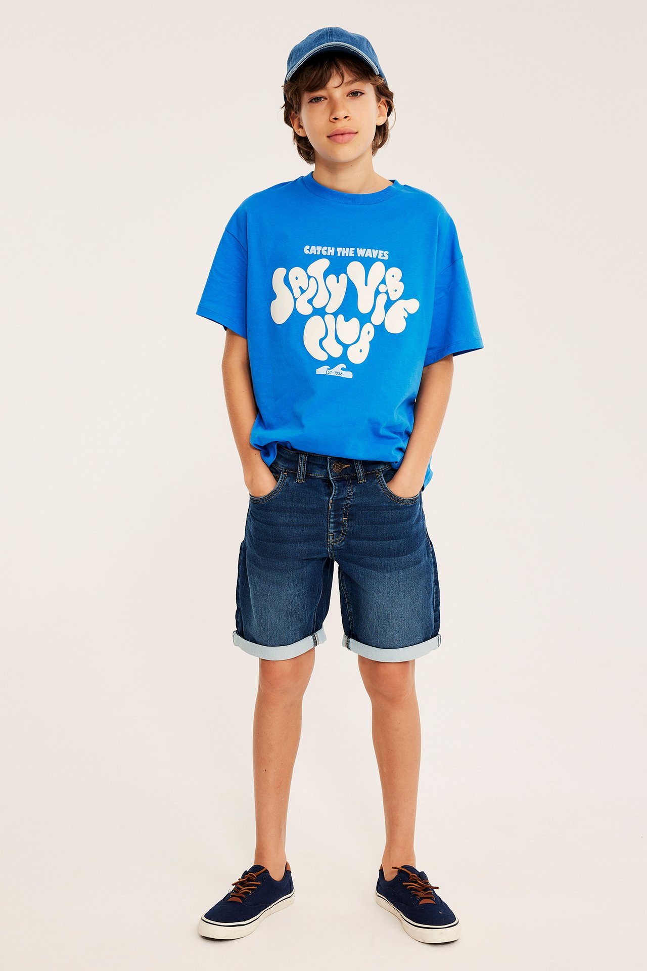 Helbild framifrån av ett barn i mörkblå jeansshorts med uppvik, blå t-shirt och blå keps.