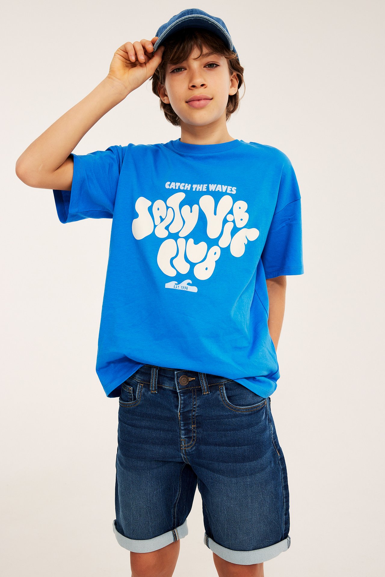 Widok z przodu dziecka w ciemnoniebieskich szortach jogger, niebieskiej koszulce z grafiką i czapce jeansowej.