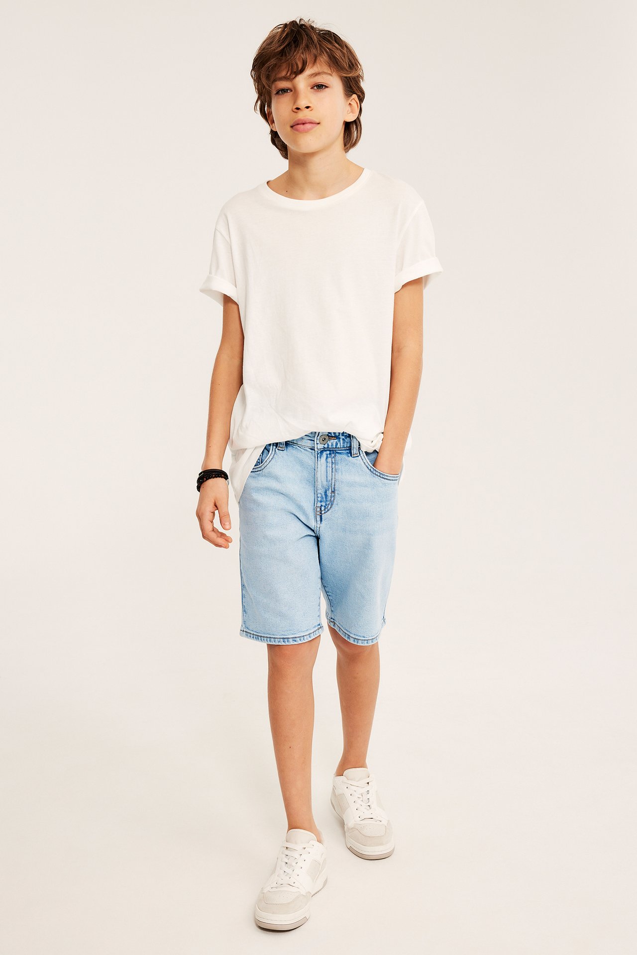 Frontvy av ett barn i ljusblå jeansshorts, vit t-shirt, vita sneakers och svart armband.