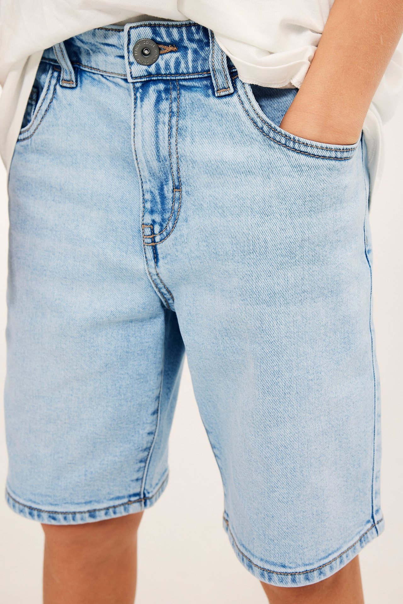 Zbliżenie: jasnoniebieskie, elastyczne szorty dżinsowe dla dziecka, zapinane na guzik, ręka w kieszeni.