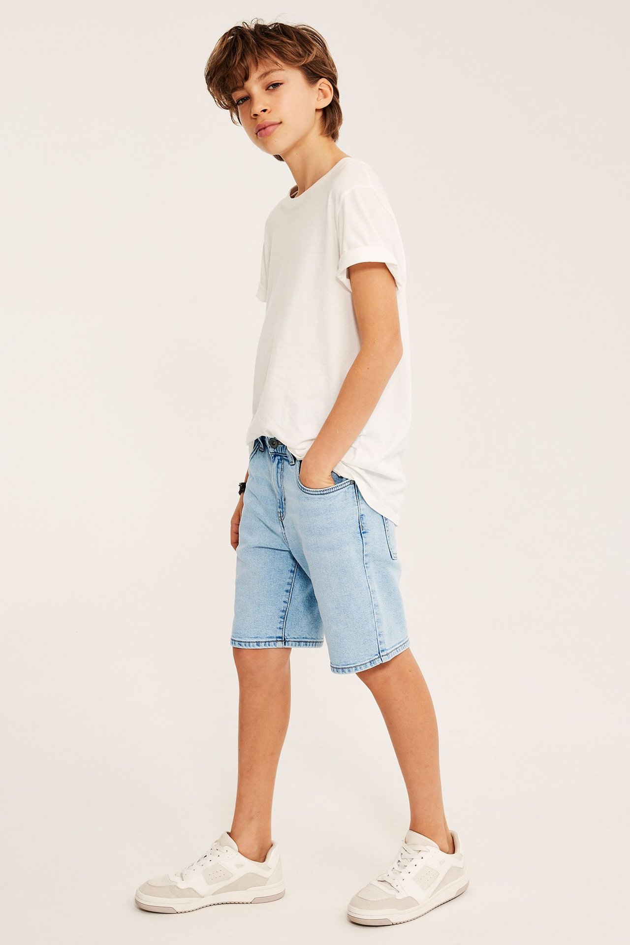 Widok z boku dziecka w jasnoniebieskich jeansowych szortach, białym T-shircie i białych tenisówkach.