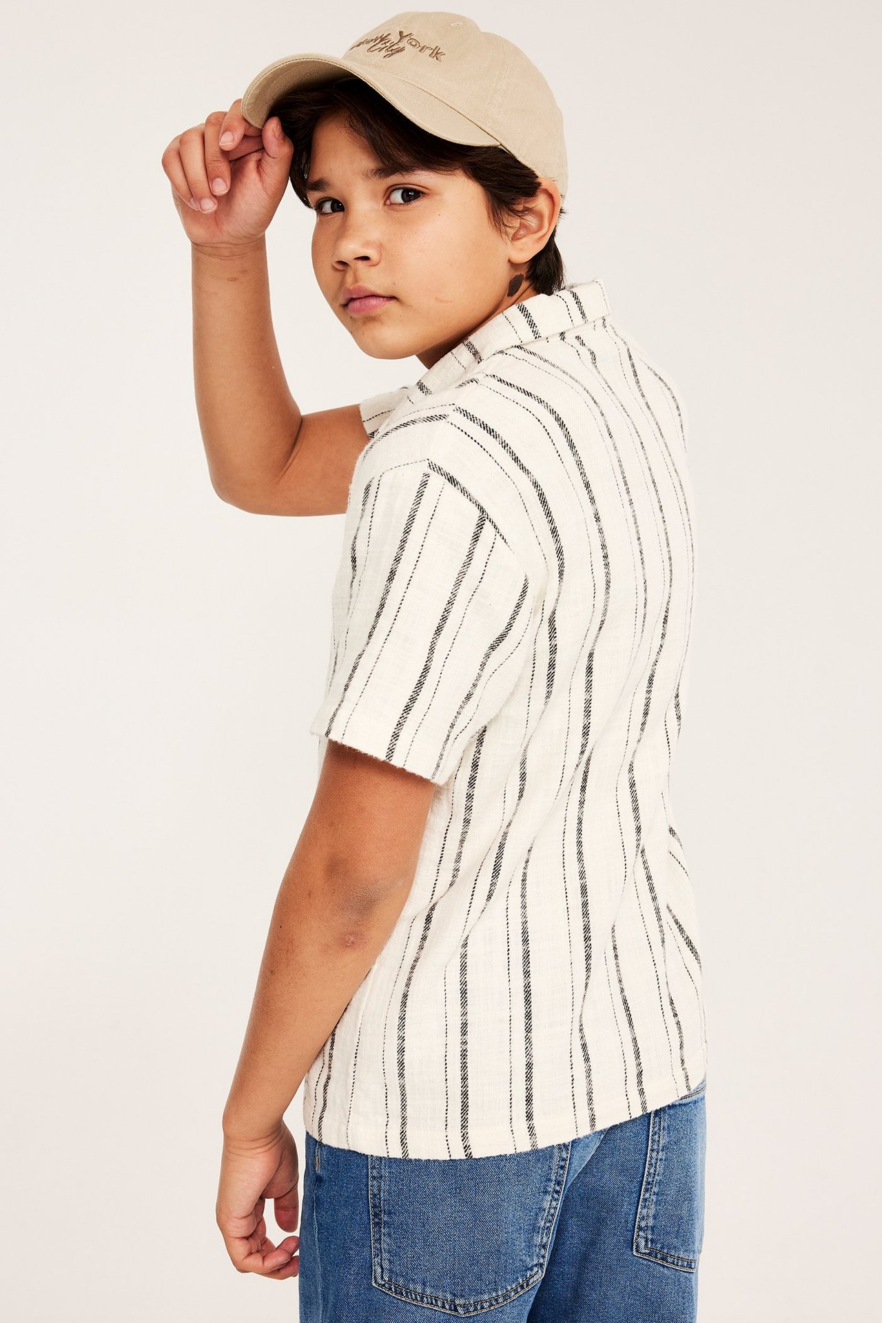 Sidevisning av et barn i offwhite stripete bomullsskjorte, blå jeans og beige caps.