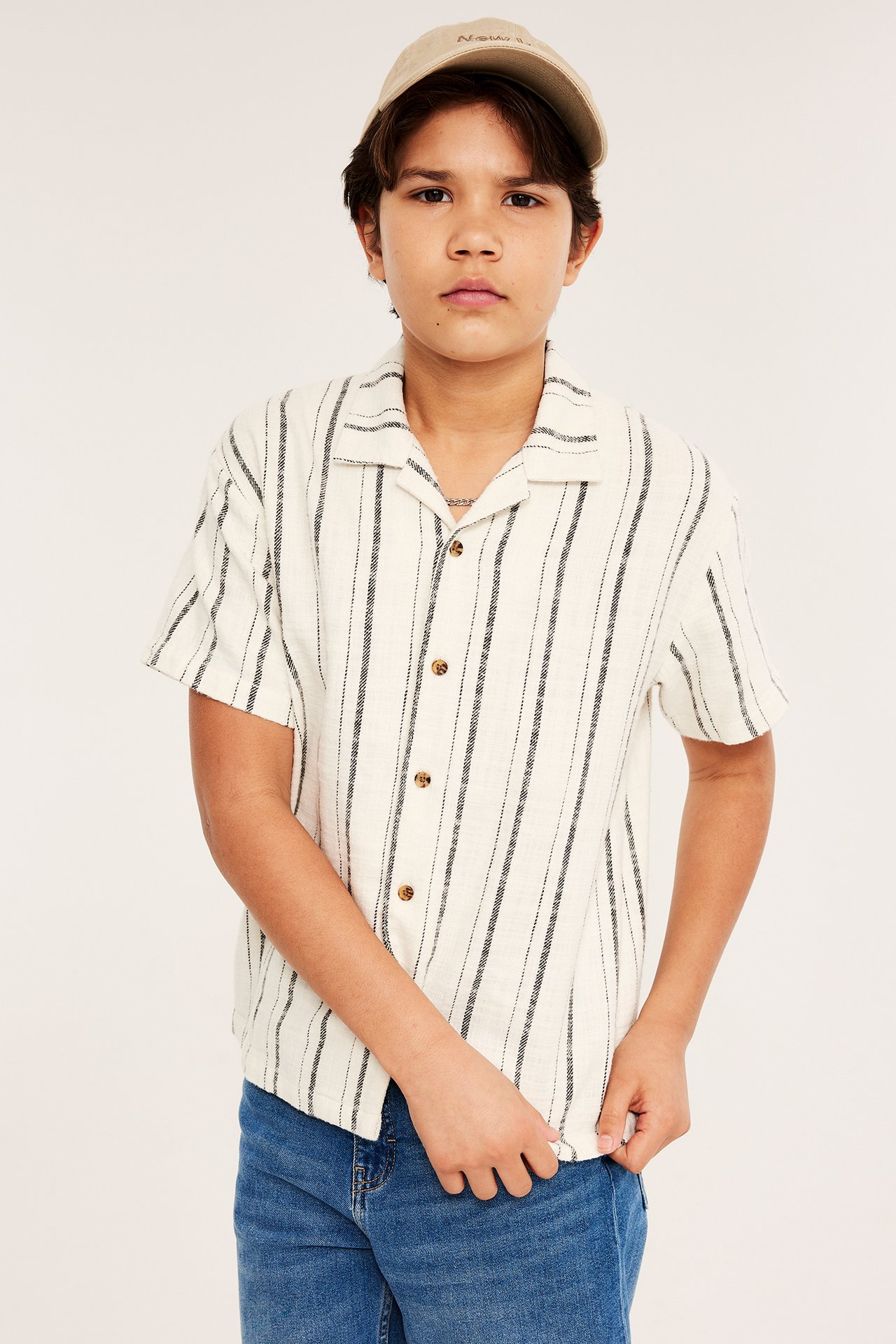 Frontbilde av et barn i en offwhite stripete bomullsskjorte, blå jeans og en beige caps.