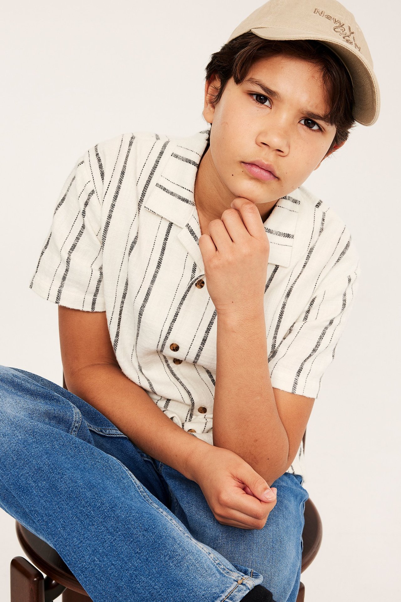 Nærbilde forfra av et barn i offwhite stripete bomulls resortskjorte, blå jeans og beige baseballcaps.