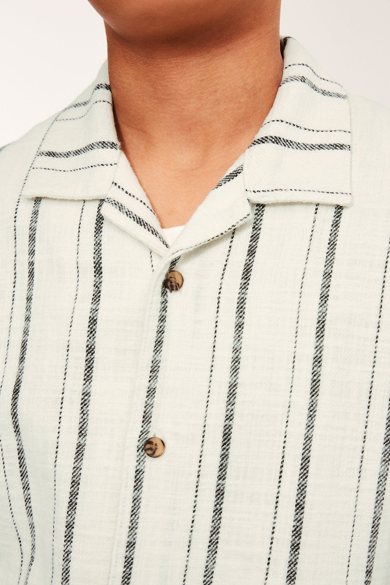 Lähikuva: Lapsi off-white raidallisessa puuvillapaidassa, ruskeat napit, valkoinen t-paita alla.