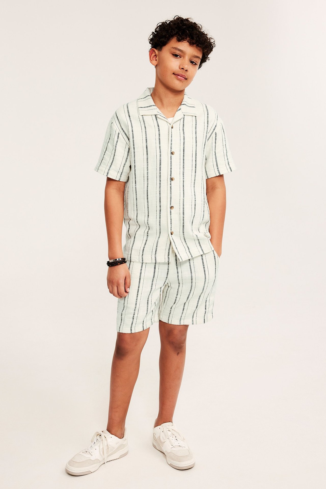 Framifrån, ett barn i off-white randig bomulls resortskjorta, matchande shorts, vita sneakers och svarta pärlarmband.