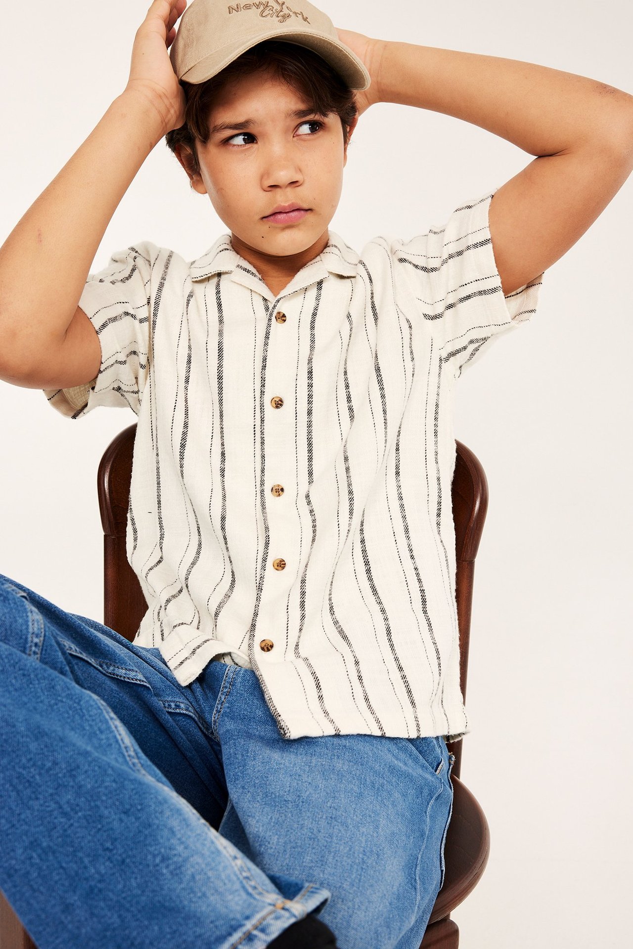 Frontbilde av et barn i en offwhite stripete bomulls-resortskjorte, blå jeans og beige caps.