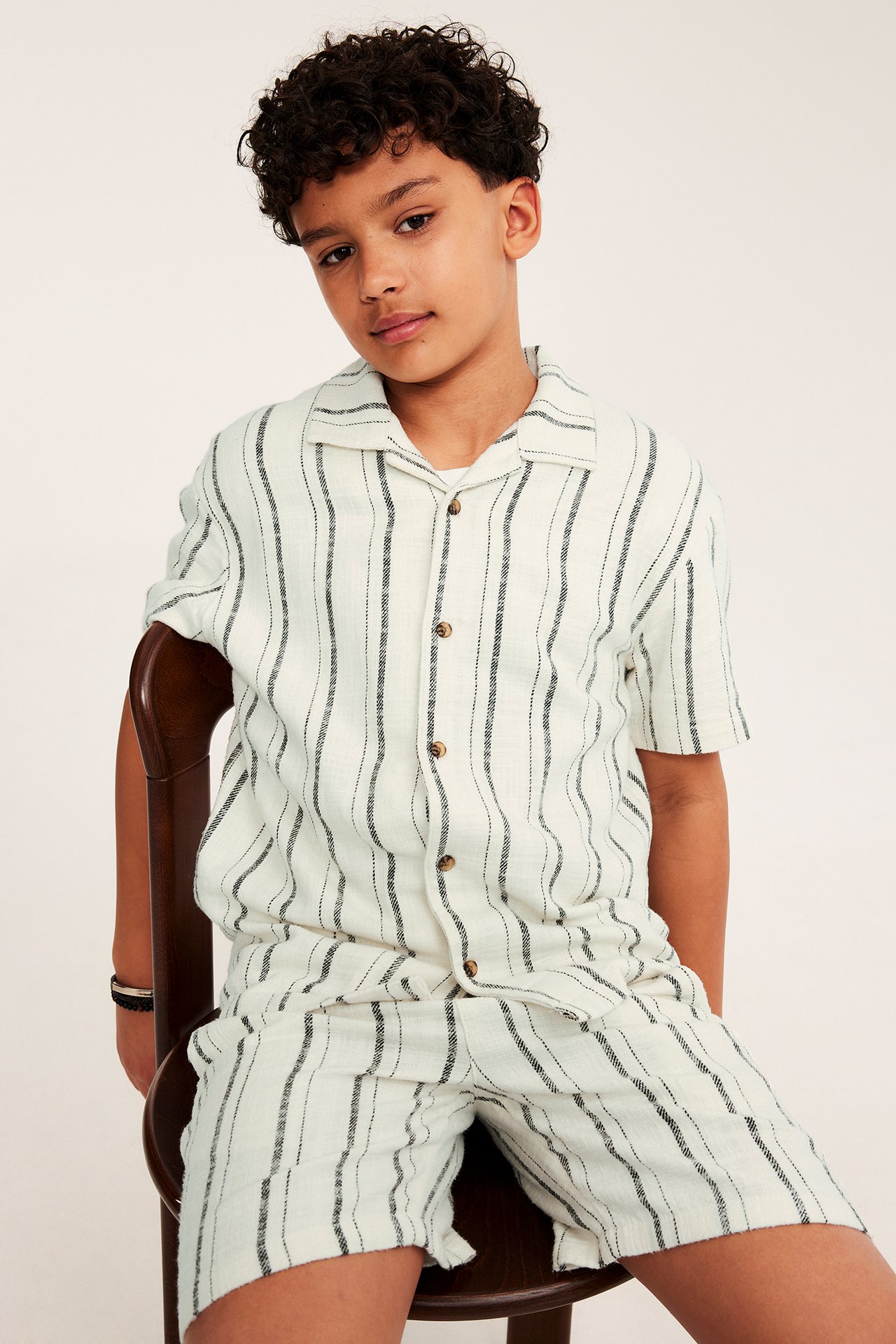 Frontbilde av et barn i en offwhite bomullsskjorte med mørke striper og matchende shorts.