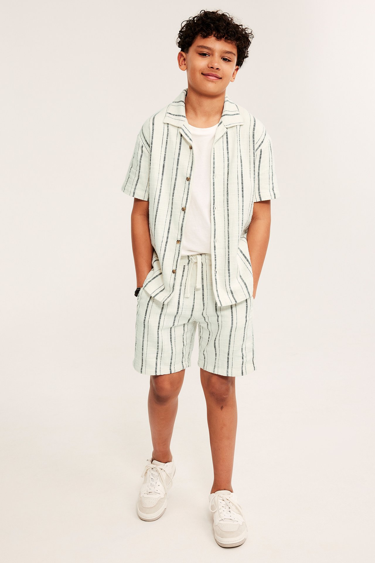 Front: Barn i offwhite stripete bomullsshorts, matchende skjorte, hvit t-skjorte og hvite joggesko.