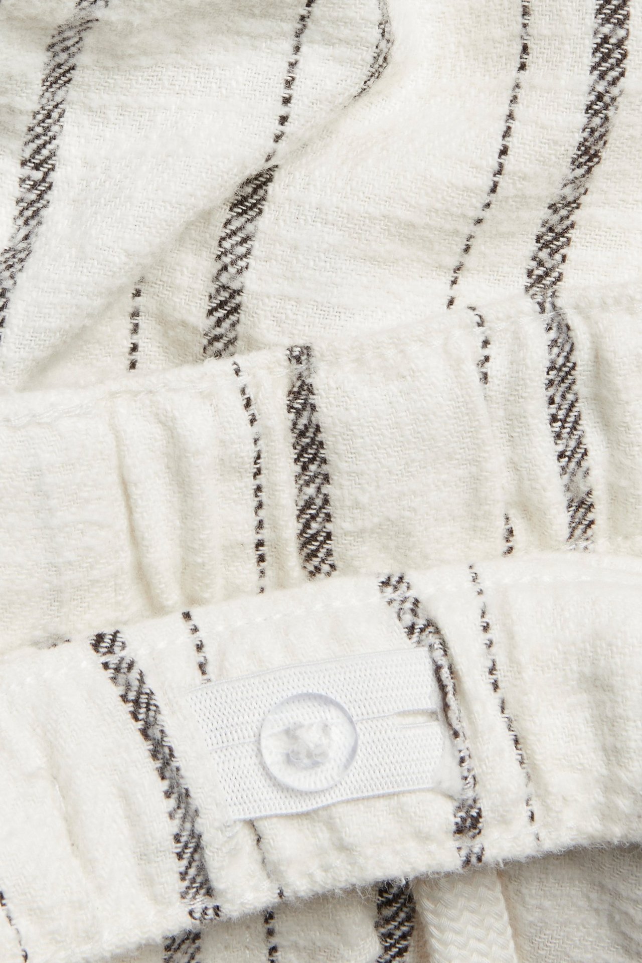 Nærbilde av offwhite stripete bomullsshorts med justerbar elastisk linning og gjennomsiktig knapp.