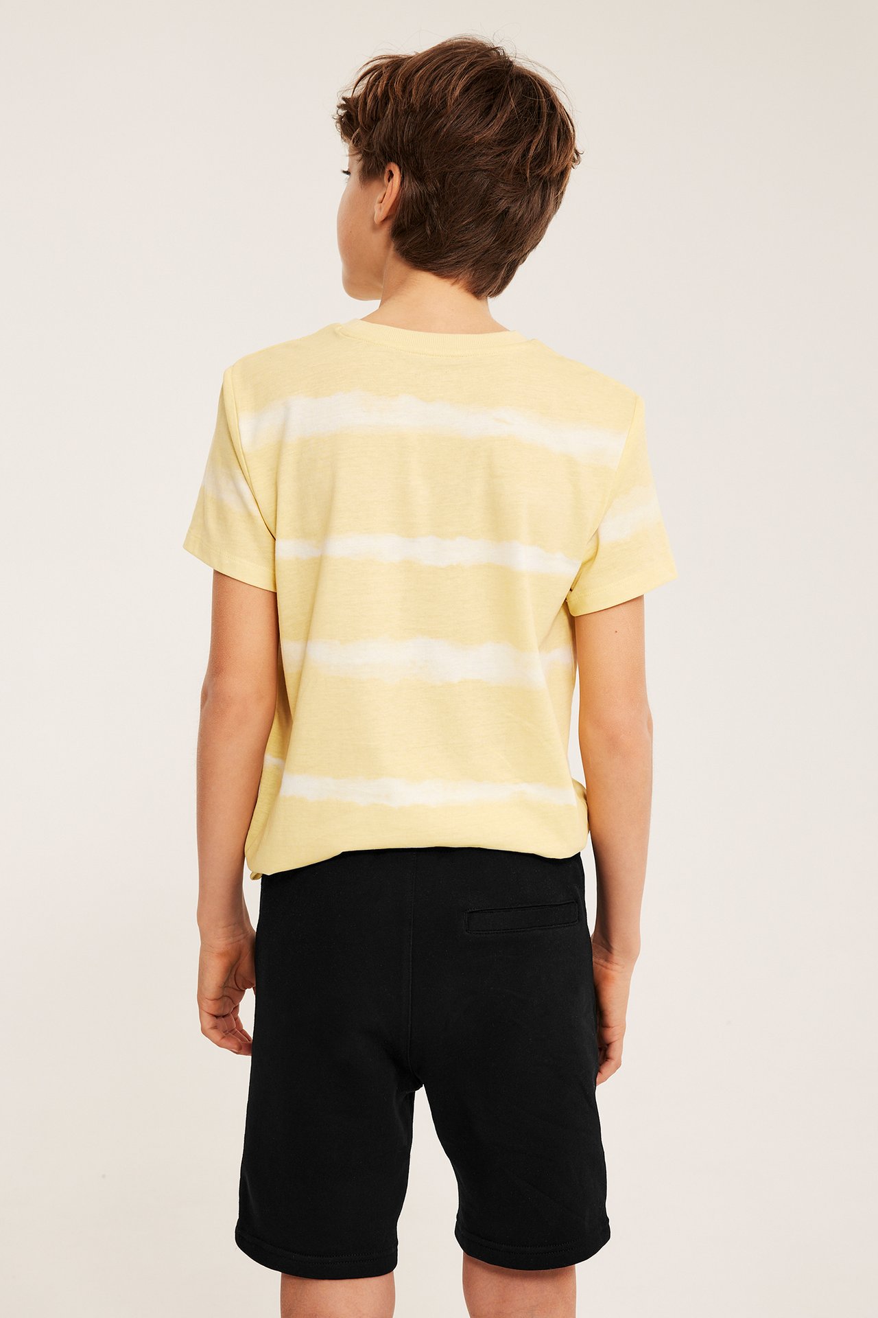 Bakfra: et barn i svarte bomulls-sweatshorts med baklomme og en gulstripet t-skjorte.