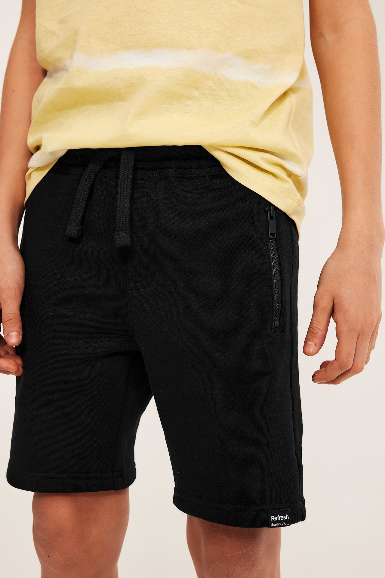 Edestäpäin: lapsi mustissa puuvillatrikoisissa college-shortseissa, joissa kiristysnyöri ja vetoketjutaskut, batikkikuvioisen t-paidan päällä.