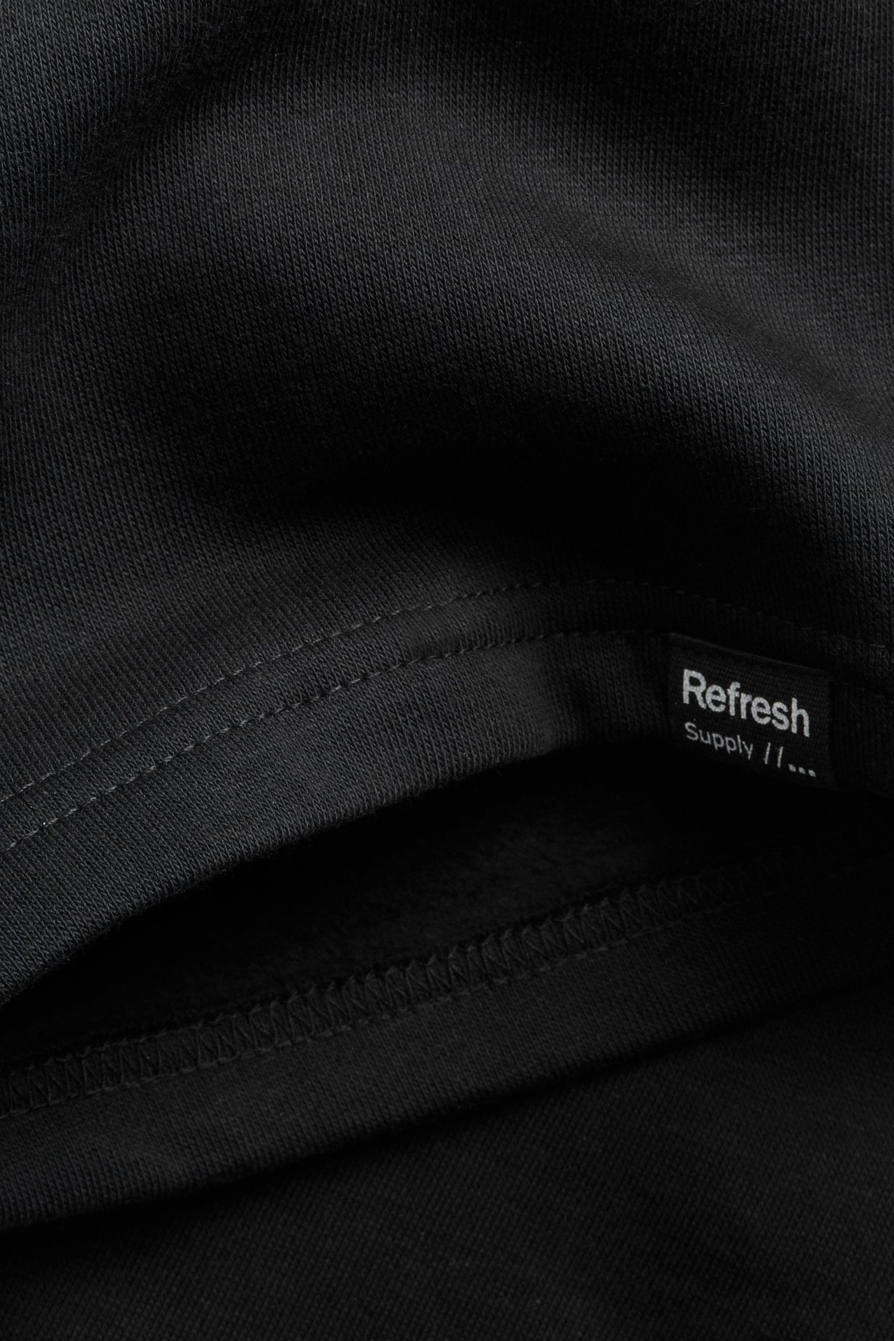 Lähikuva mustasta puuvillatrikooshortsikankaasta, jossa näkyvä tikkaus ja pieni Refresh Supply -etiketti.