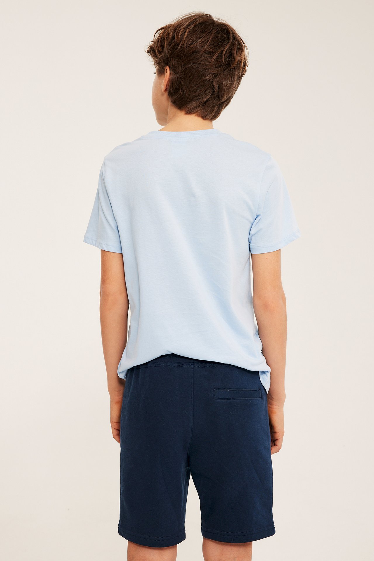 Barn, bakfra: mørkeblå bomulls-sweatshorts med baklomme og en lys blå t-skjorte.