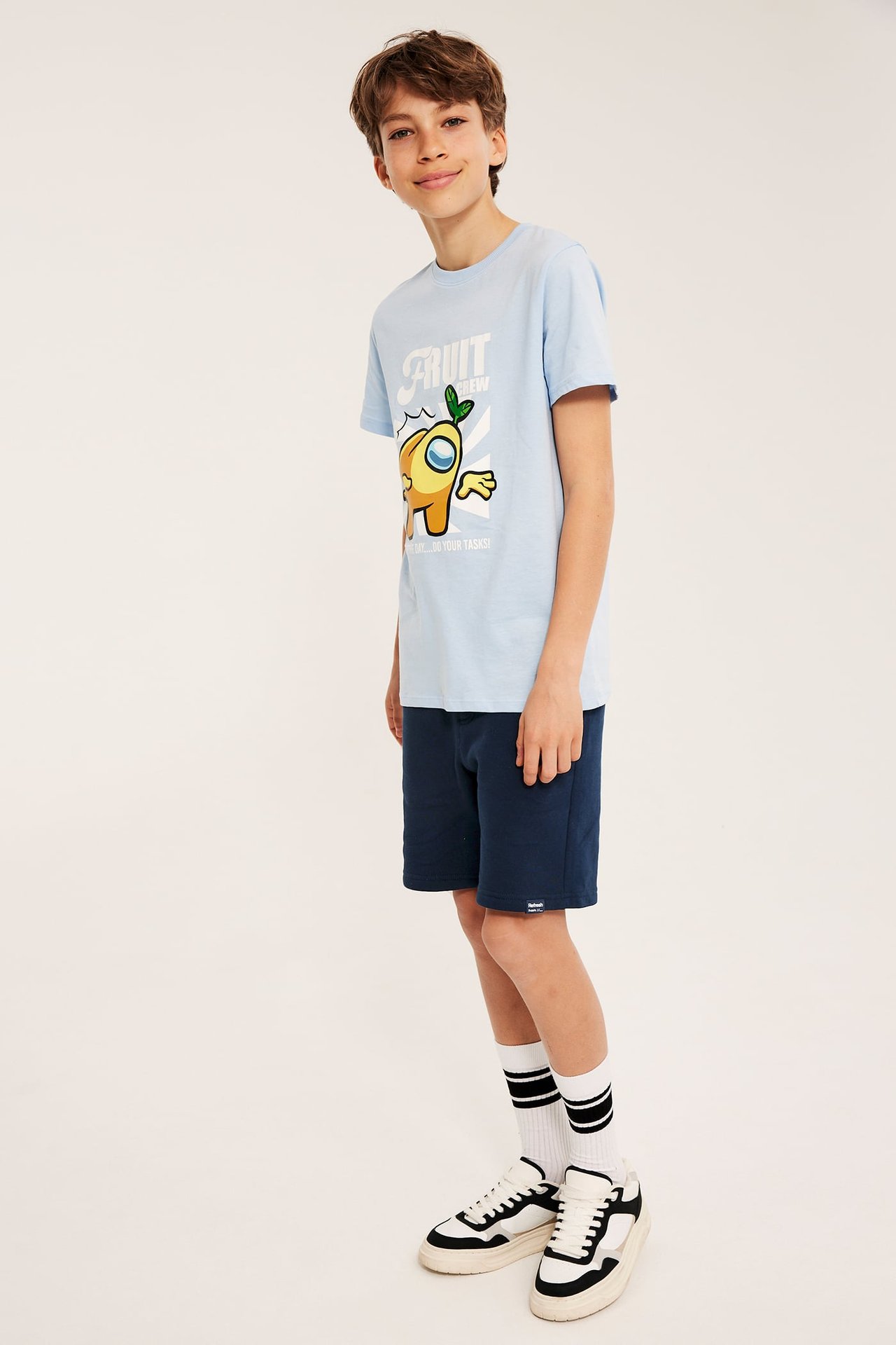 Sidenfra-frontbilde av et barn i mørkeblå bomulls-sweatshorts, lys blå t-skjorte, stripete sokker, sneakers.